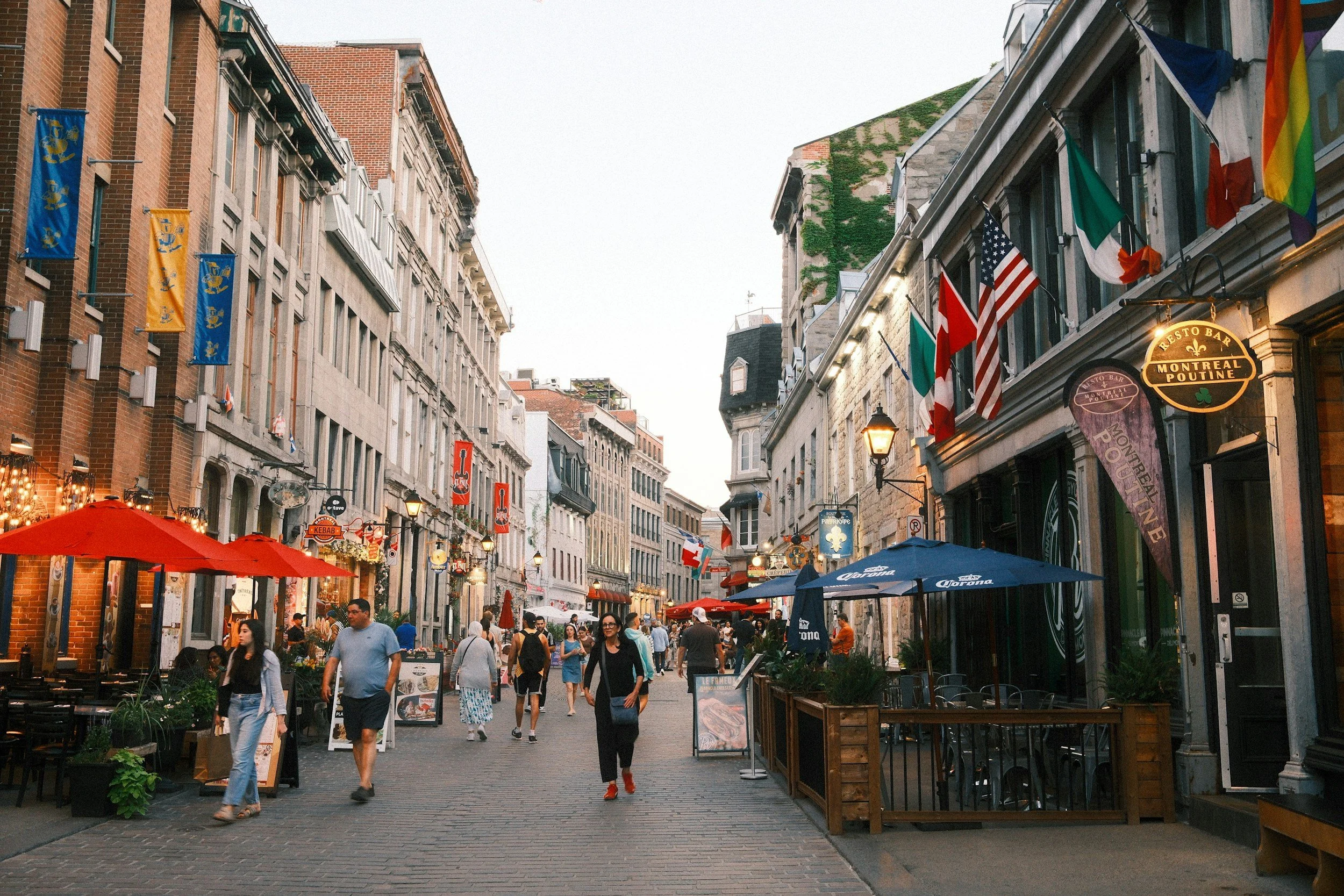 A busy pedestrian street in Montreal with outdoor dining, flags, and umbrellas, with people walking and shops on both sides.