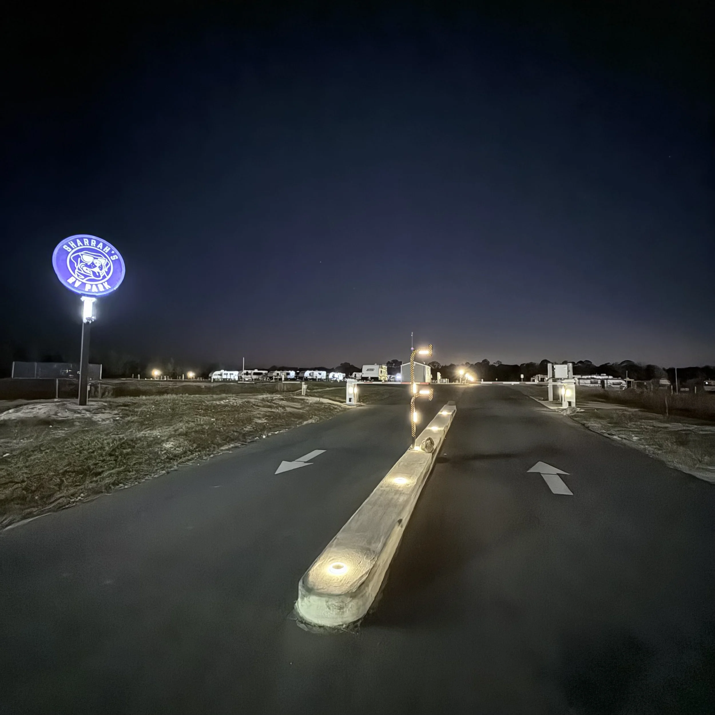 Nighttime scene at a vehicle park with a sign that says 'WARRAH'S RV PARK'. The entrance has a narrow road with a concrete median barrier, illuminated with small lights. There are parked RVs and trailers in the background under a dark sky.