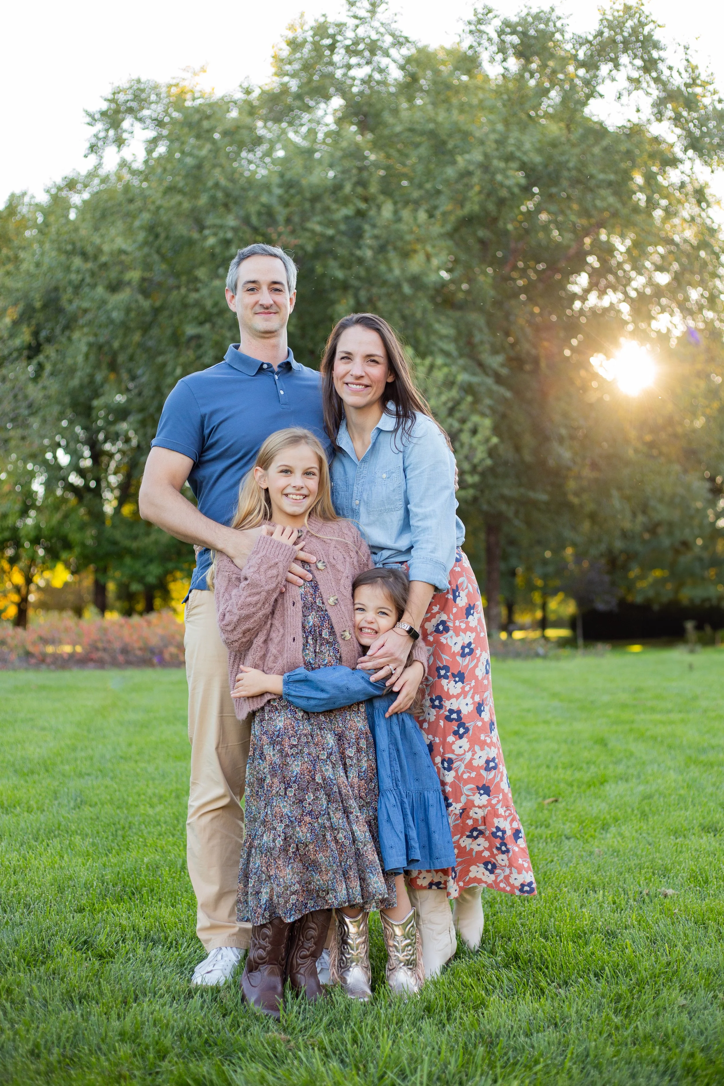 A family of four standing on a grassy field in a park during sunset. The parents are behind their two daughters, smiling at the camera.