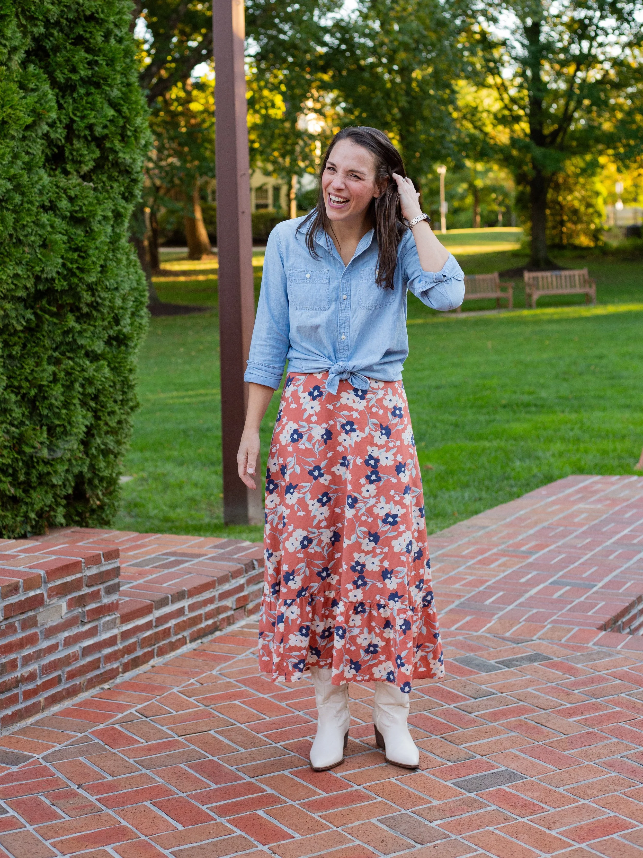 A woman with dark brown hair, wearing a light blue denim shirt tied at the waist, a long floral skirt, and white cowboy boots, standing outdoors on a brick pathway, smiling and touching her hair in a park with green grass, trees, and benches in the background, during daytime.