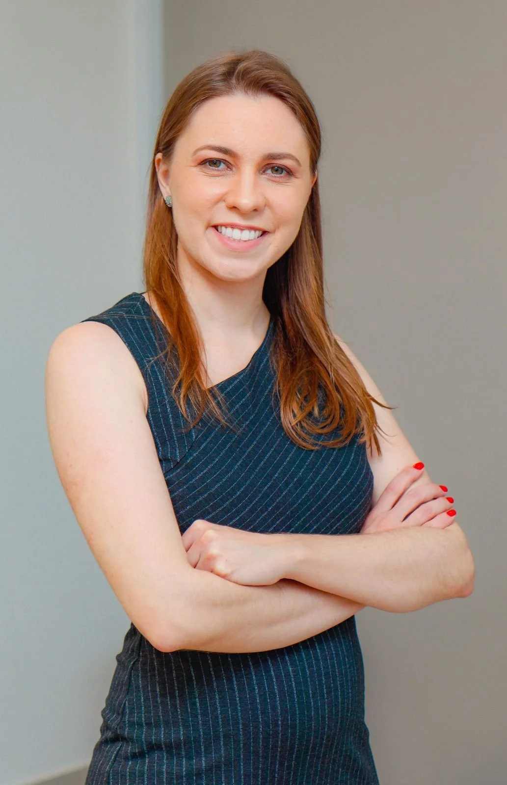A woman with red hair, blue eyes, and fair skin smiling, wearing a sleeveless dark blue dress with thin vertical stripes, standing with her arms crossed in front of a plain wall.
