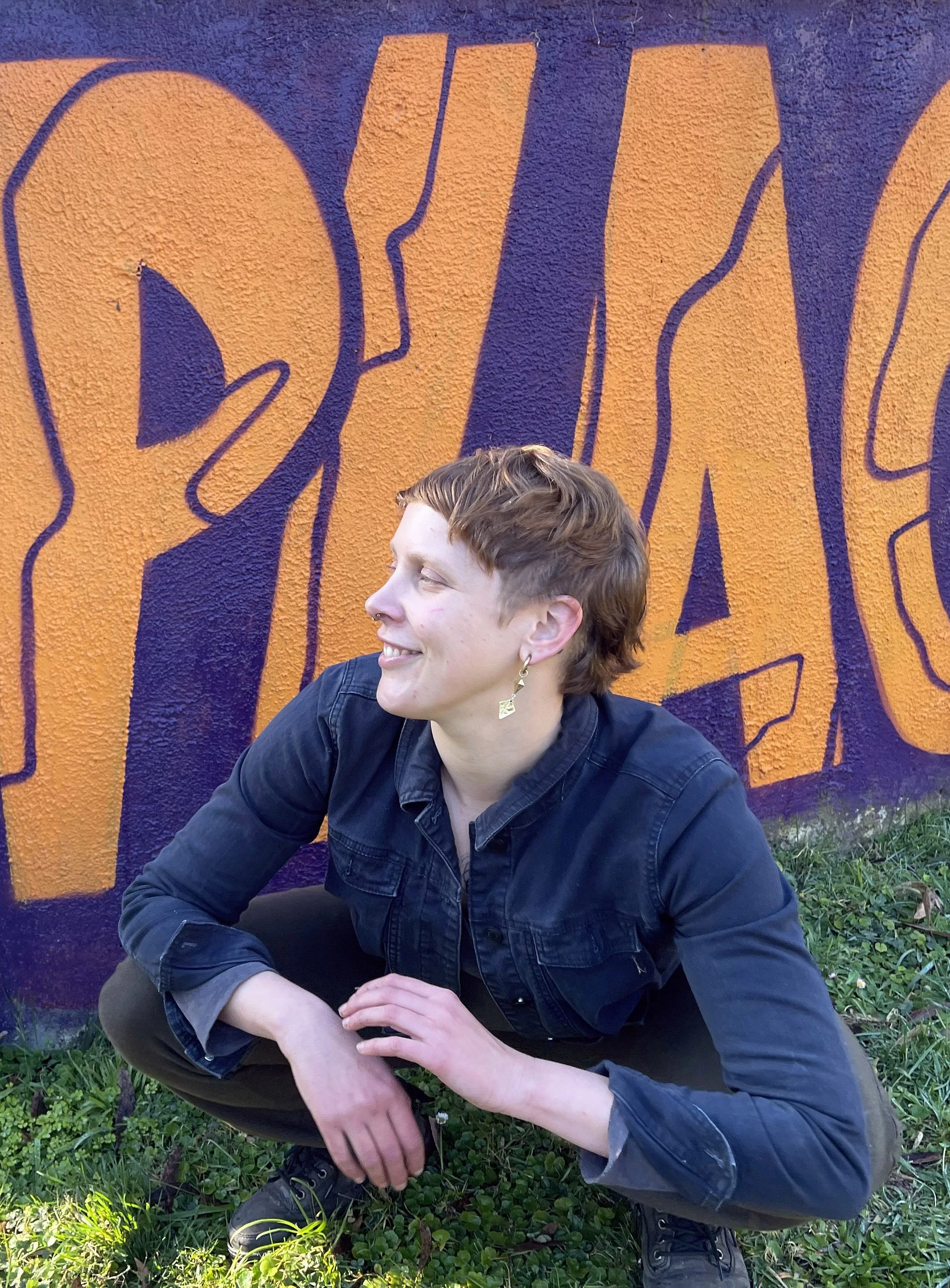 A woman with short reddish hair crouching on grass in front of a brightly colored graffiti wall that features orange and purple lettering.