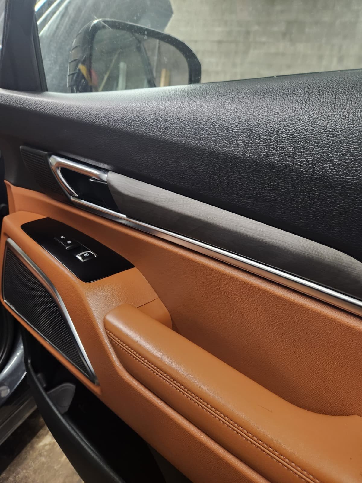 Close-up of a car door interior with black, gray, and tan leather materials, silver door handle, and window control buttons.