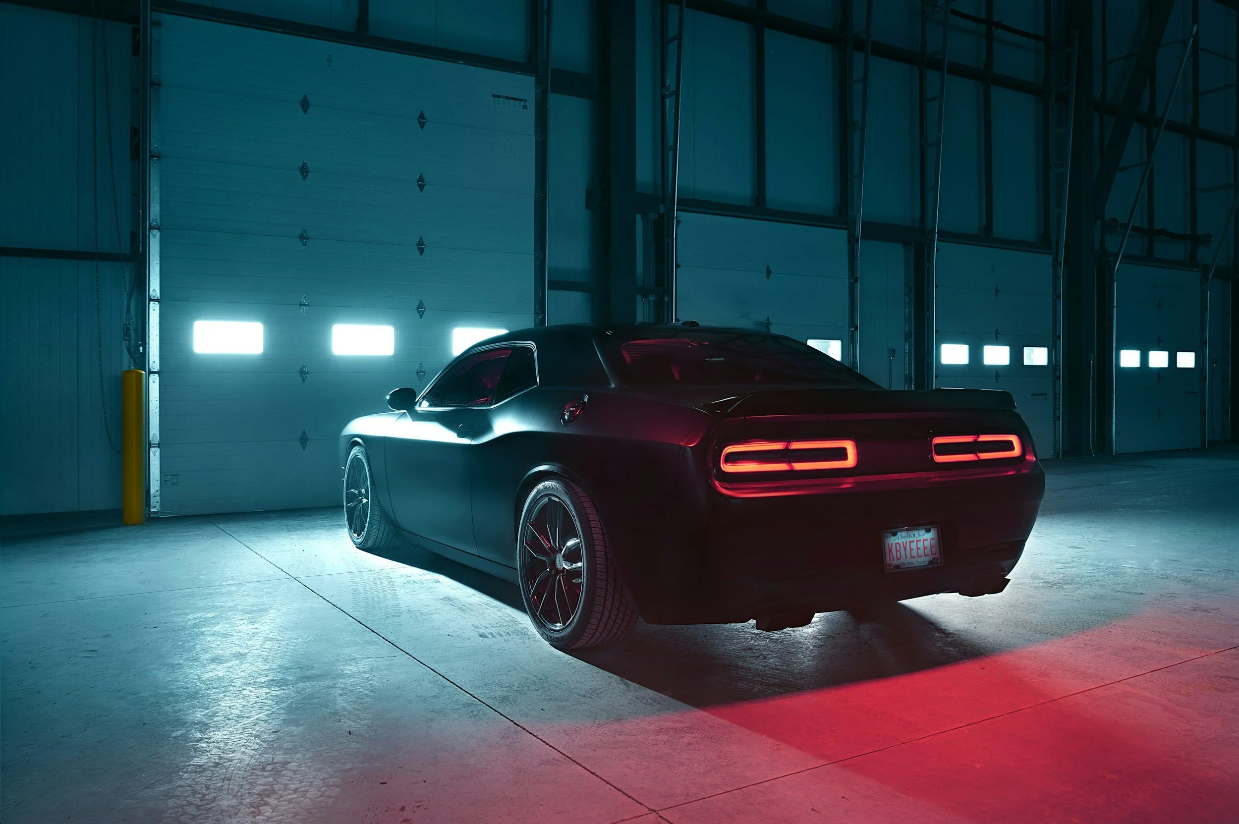 A black sports car with red tail lights parked in a dimly lit warehouse with large garage doors and blue lighting.