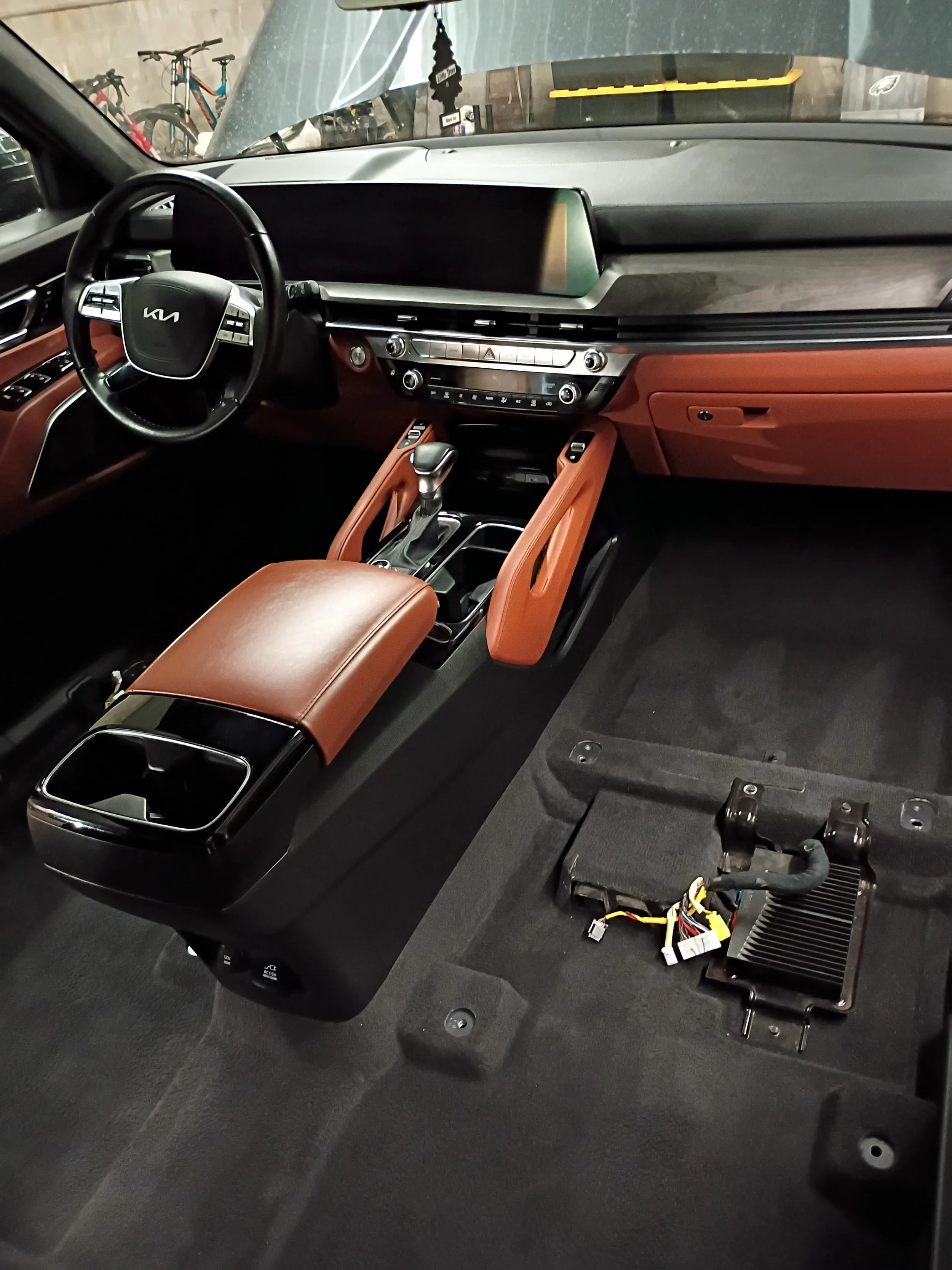 The interior of a modern Kia vehicle with a tan dashboard and black accents, including a steering wheel, digital display, and center console. The passenger side floor is exposed, revealing wiring and electronic components.