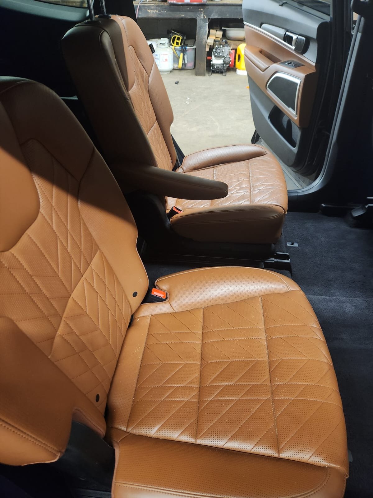 Tan leather vehicle seats inside a car with the passenger door open, showing a garage background with tools and equipment.