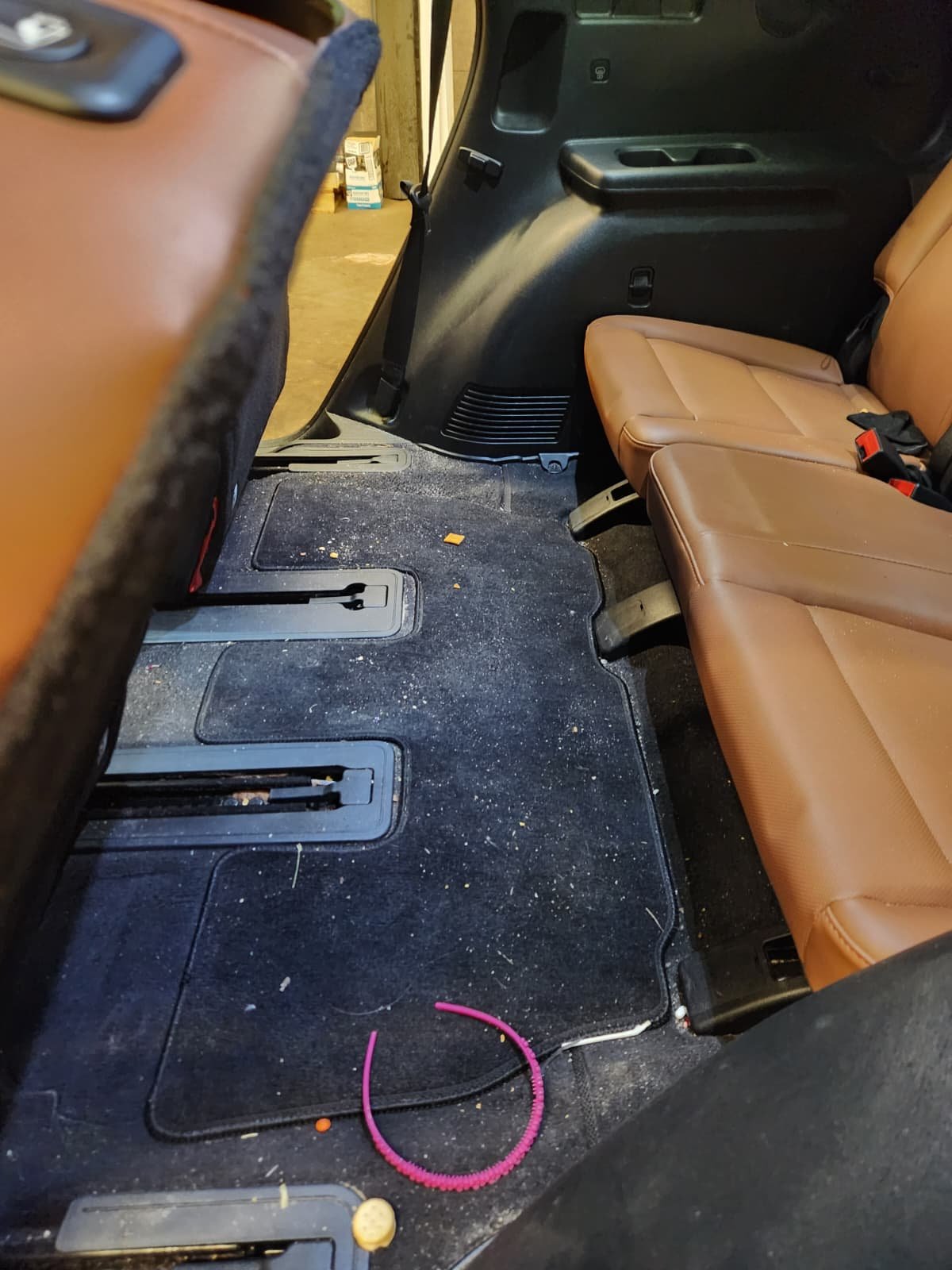 Backseat of a vehicle showing dirty black carpet with crumbs and debris, tan leather seats, and various small objects including a pink spiral cable and a yellow item on the floor.