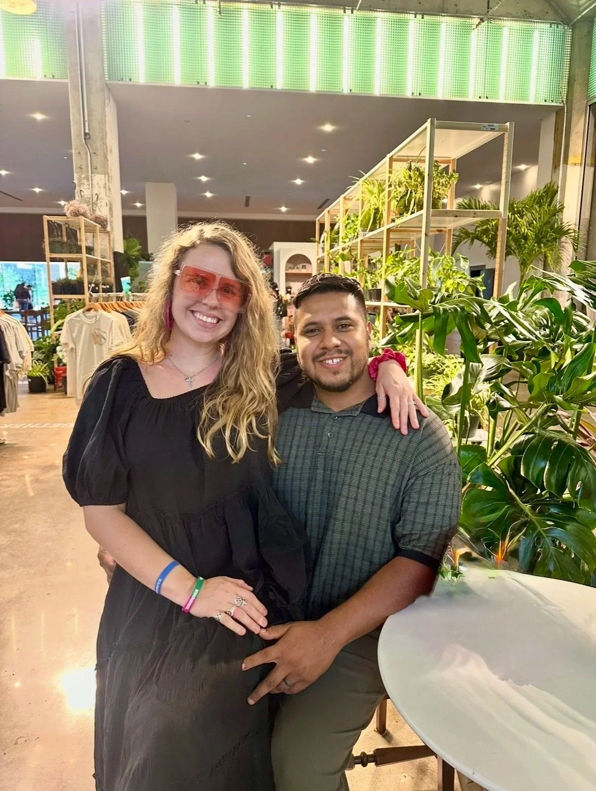 A smiling woman with blonde hair, pink sunglasses, wearing a black dress, standing next to a man in a green plaid shirt, with their arms around each other, in a bright, indoor plant-filled setting.