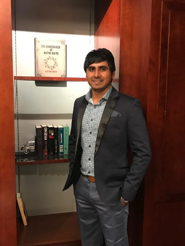 A man in a tuxedo standing in front of a bookshelf with books and decorative items.