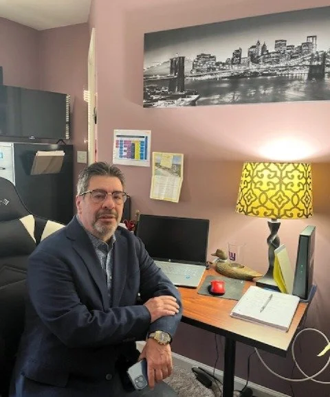 Man in a suit sitting at a desk with a computer, laptop, and notebook in a home office, decorated with a cityscape photo and a yellow patterned lampshade.