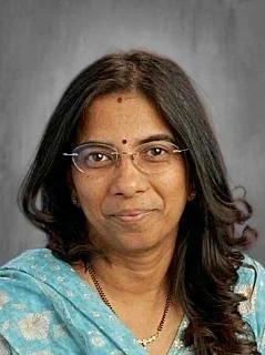 A woman with long black hair wearing a sleeveless, light-colored dress stands in front of a green shrub and a mirror in an indoor setting.