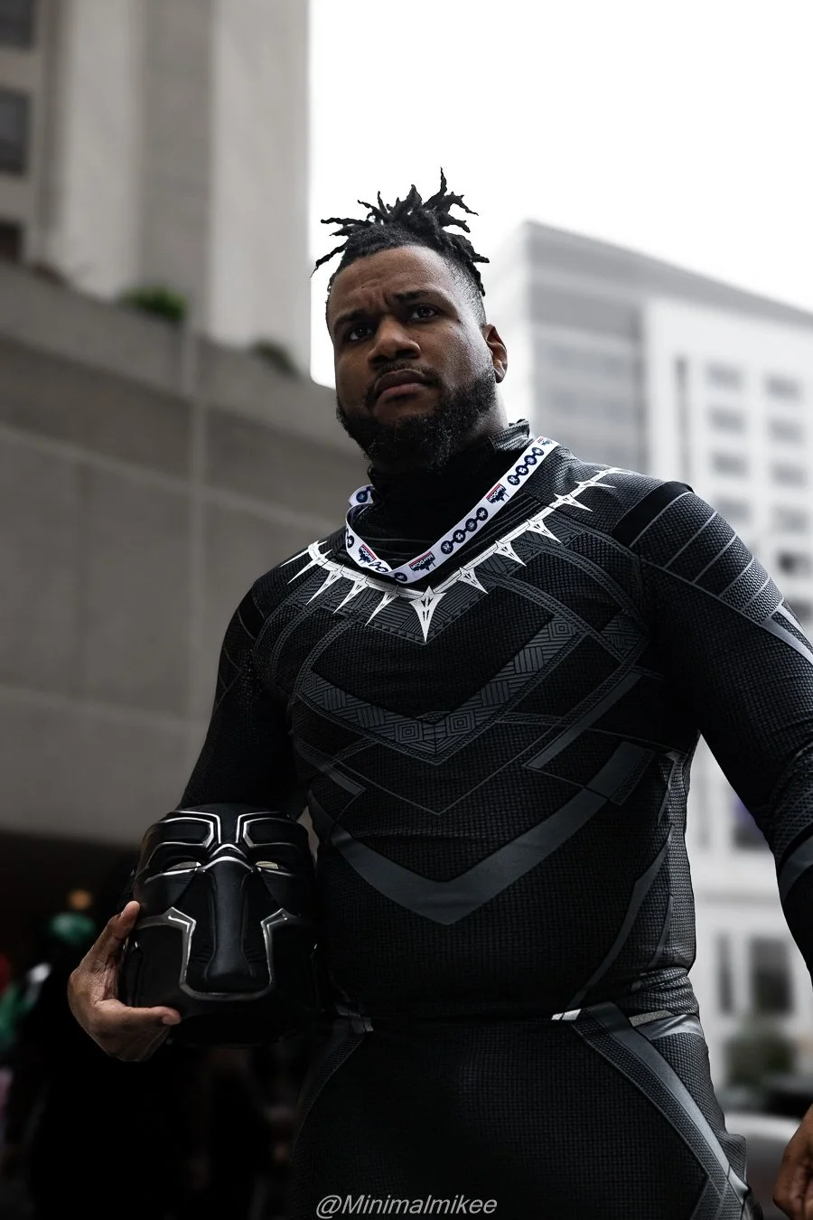 A man dressed in Black Panther-themed Black Panther costume from Marvel, holding the Black Panther mask, standing outdoors in an urban area with tall buildings in the background.