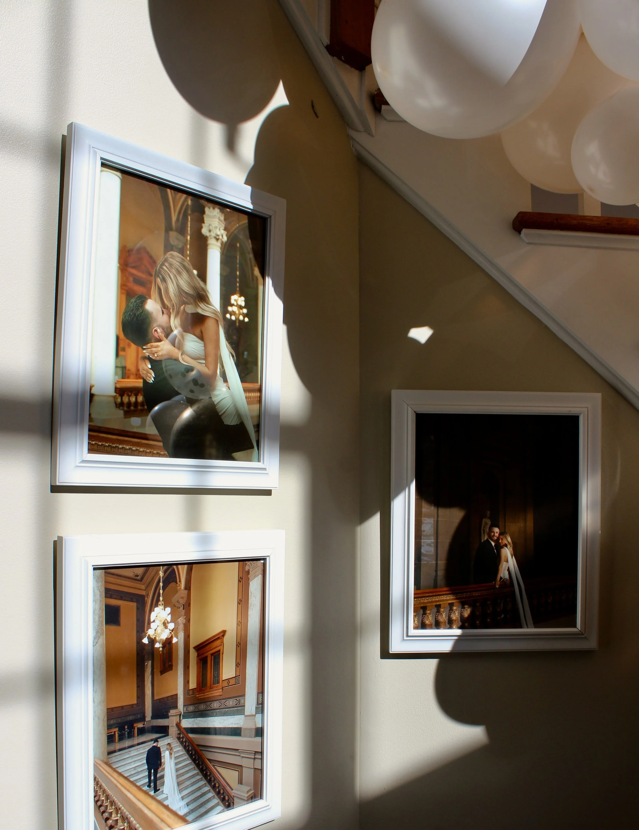 A series of framed photographs on a wall, with the main photo showing a couple kissing on a staircase in an elegant, historic building.