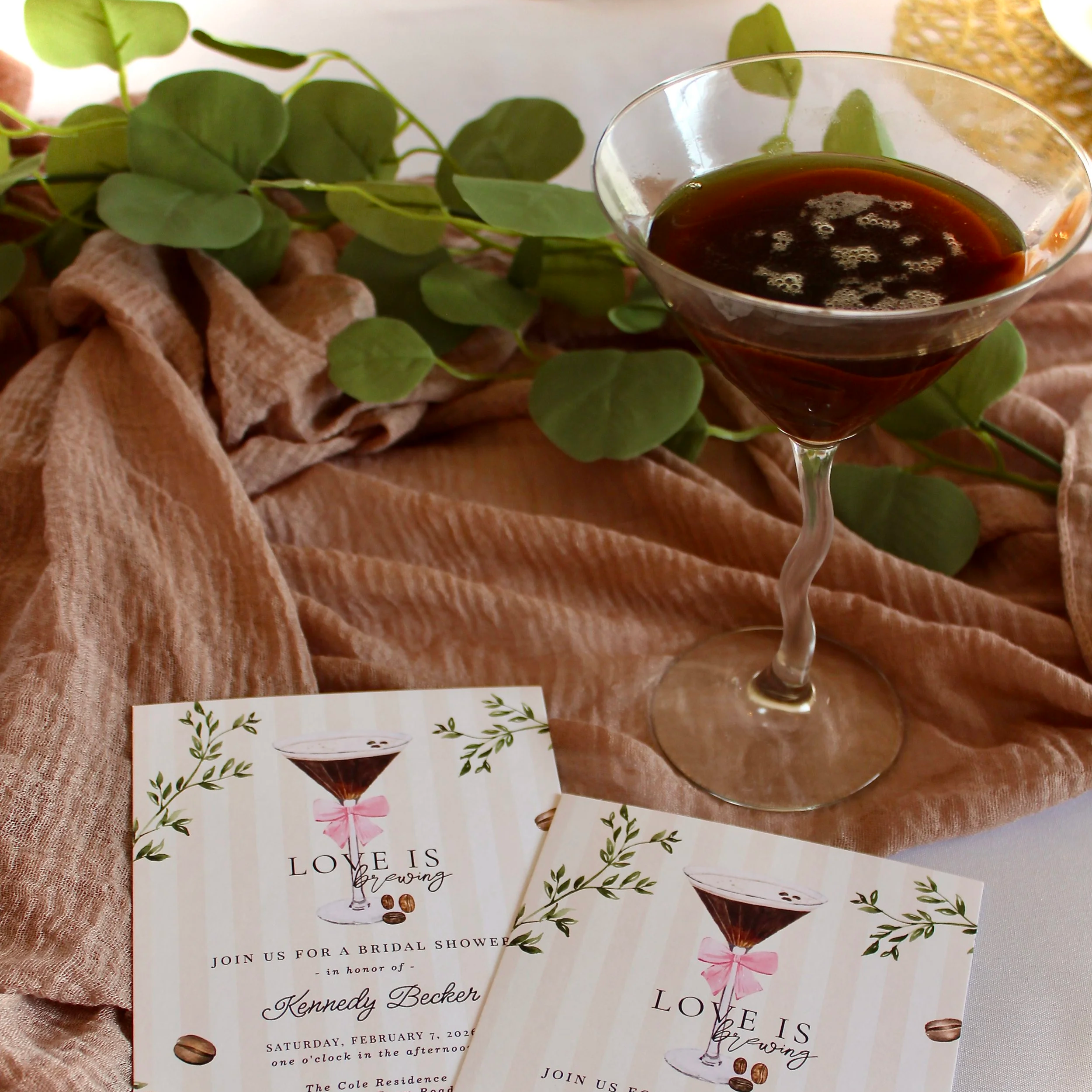 A bridal shower invitation featuring a glass of dark coffee or tea, green leaves, a pink ribbon, and coffee beans on a peach-colored fabric background.