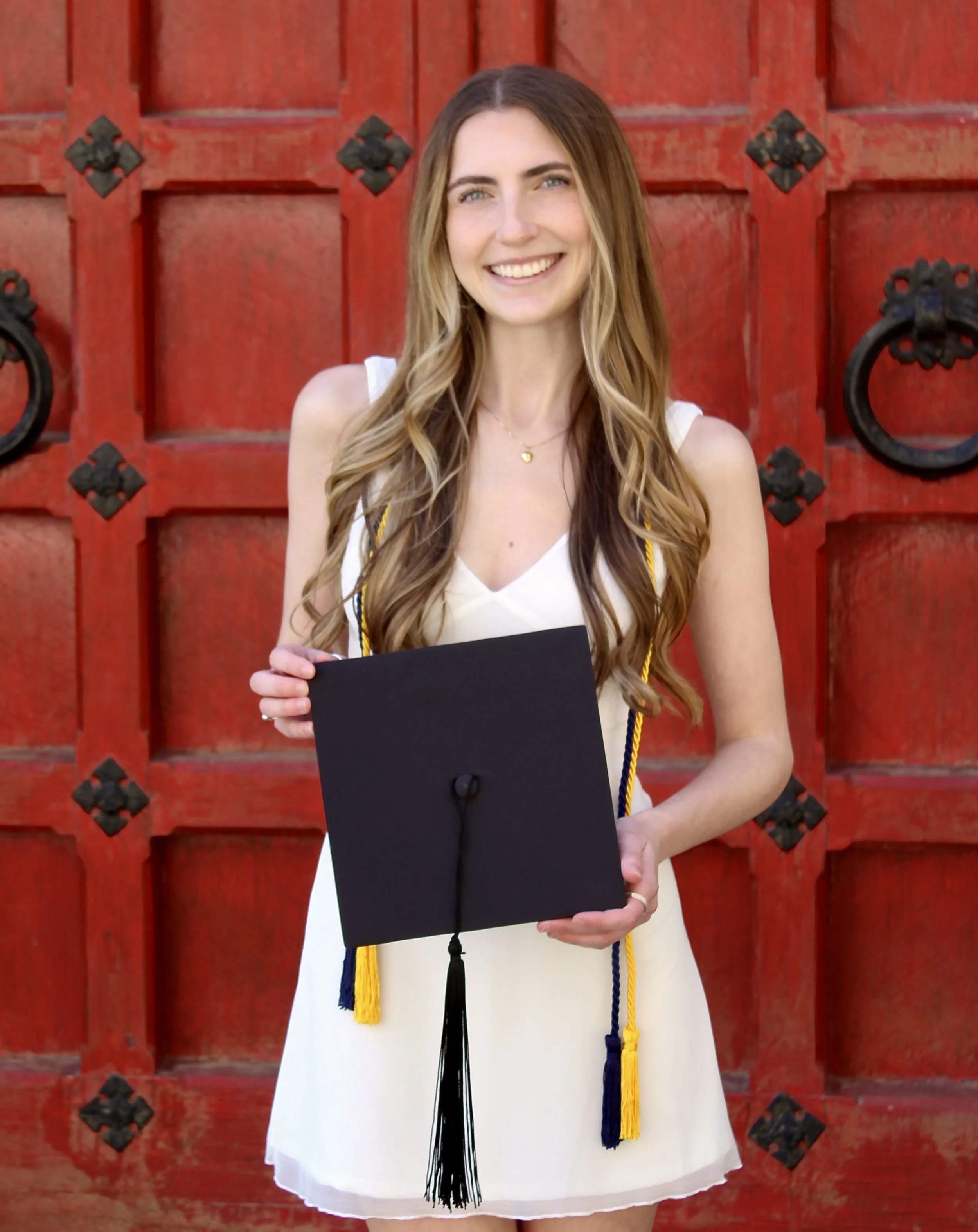 A young woman with long, wavy hair wearing a white dress and holding a graduation cap, standing in front of a red wooden door with black wrought iron accents.