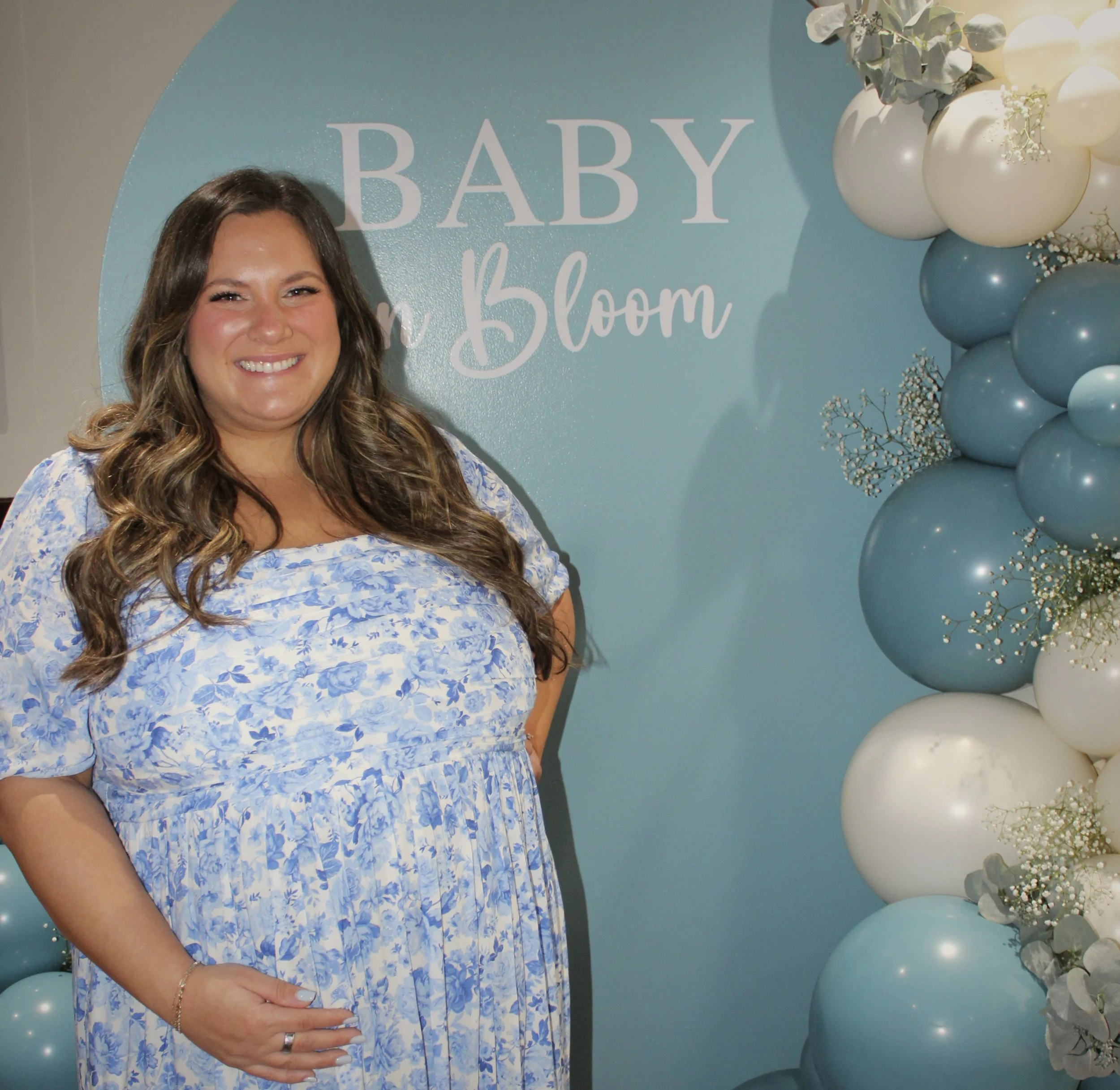 A woman in a floral blue and white dress standing in front of a blue backdrop with the words 'Baby in Bloom' and a balloon decoration of white, blue, and gray balloons with sprigs of baby's breath and greenery.