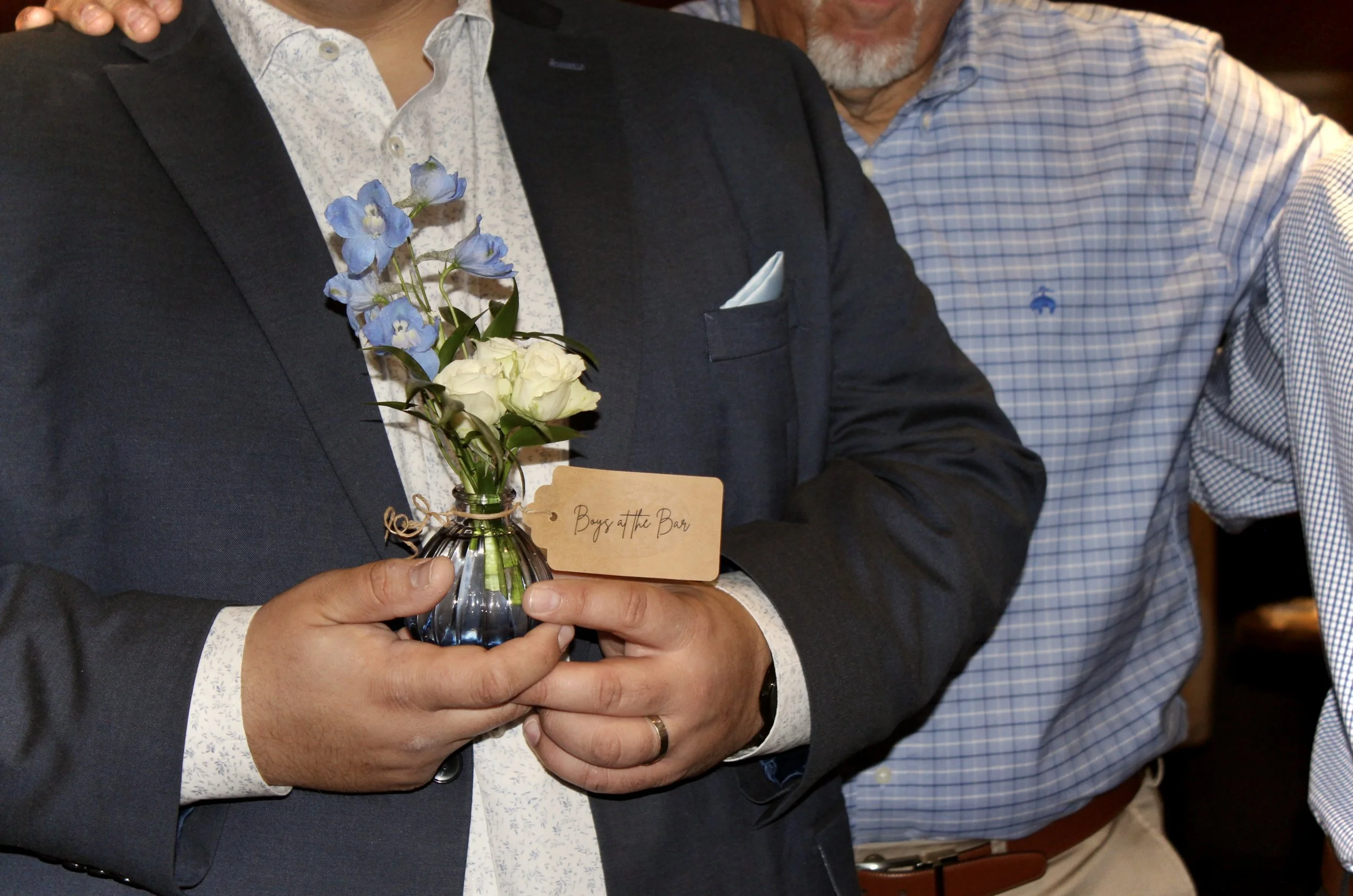 Three men dressed in suits are standing close together at an event, with one of them holding a small vase with blue delphinium and white roses, along with a card that reads 'Boys at the Bar'.