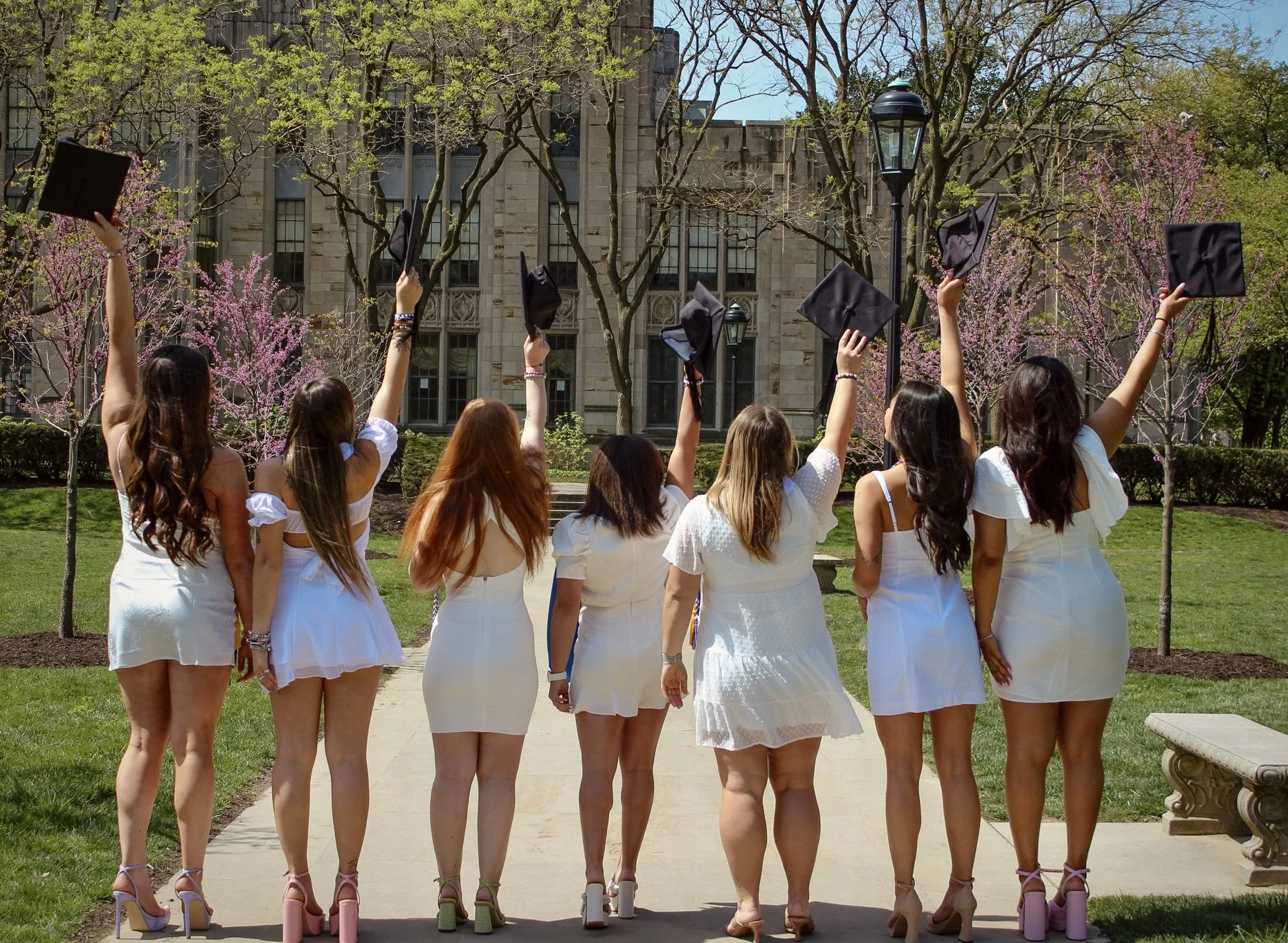 A group of young women dressed in white dresses standing outdoors, raising their graduation caps in the air, with a historic building and trees with pink blossoms in the background.