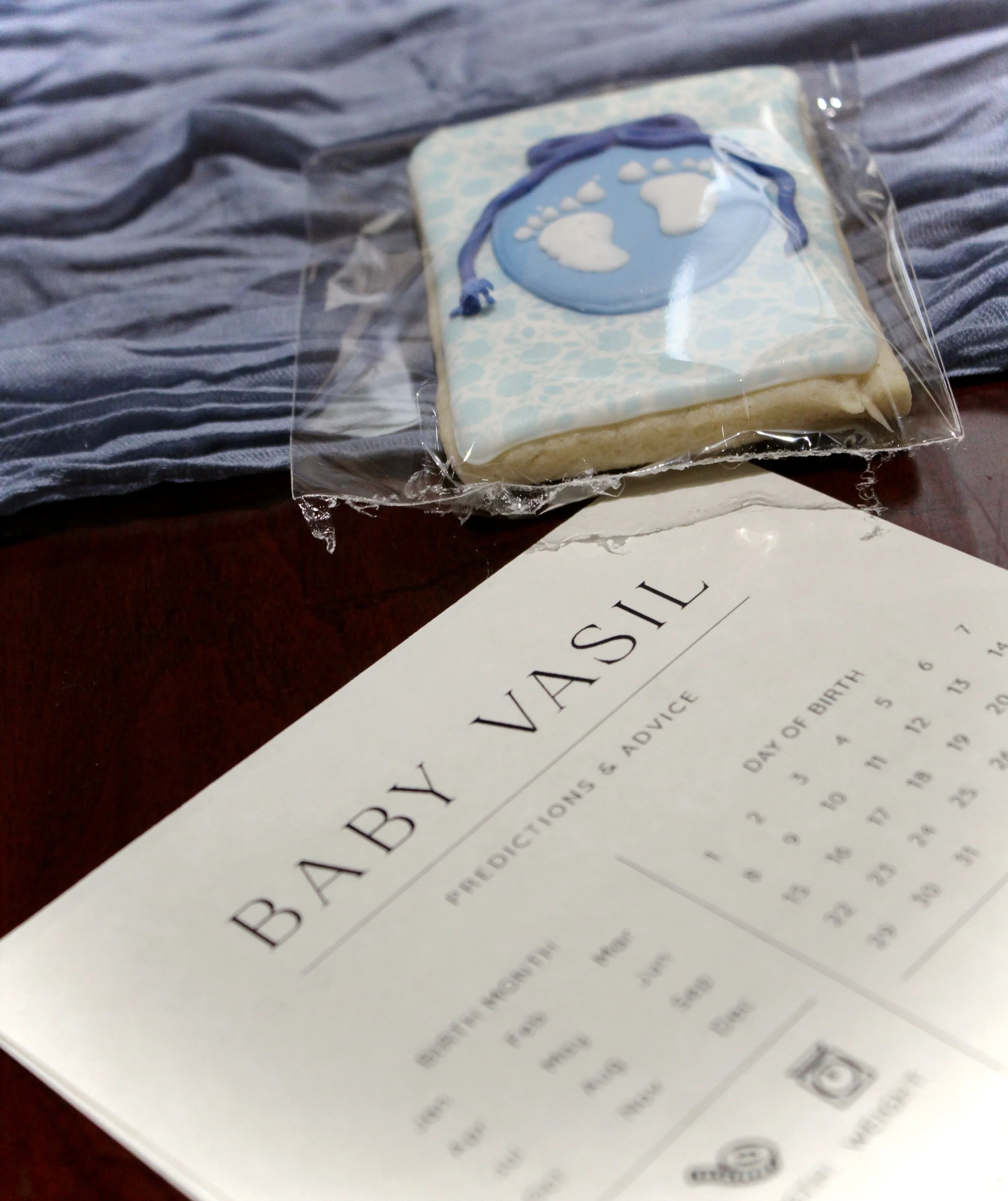 A decorated sugar cookie with a snowy design, featuring blue and white icing with footprints and a blue ribbon, inside a clear plastic bag, resting on a letter with typed text.