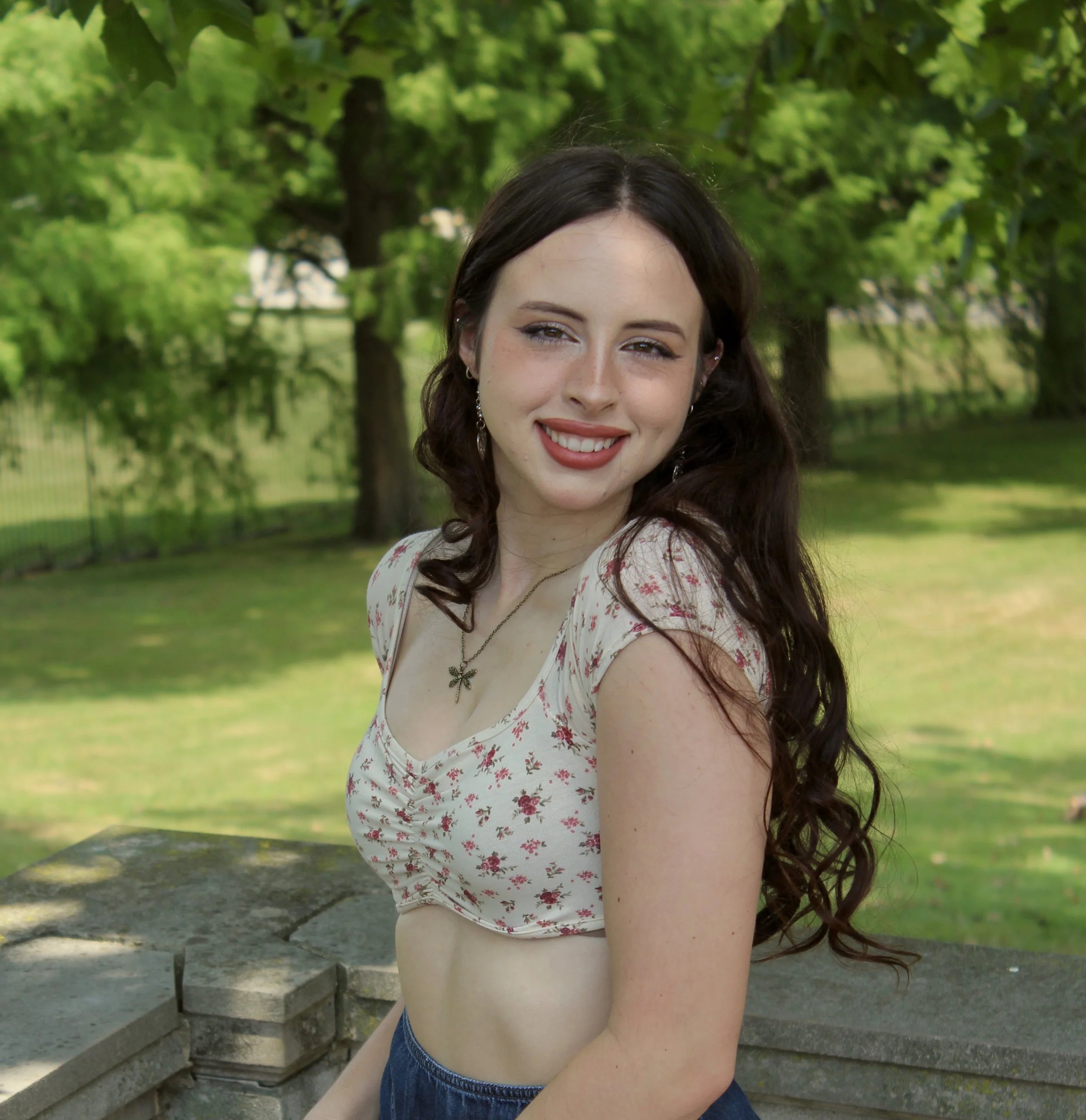 A young woman with dark brown hair and freckles smiling outdoors in a park with lush green trees and stone steps.