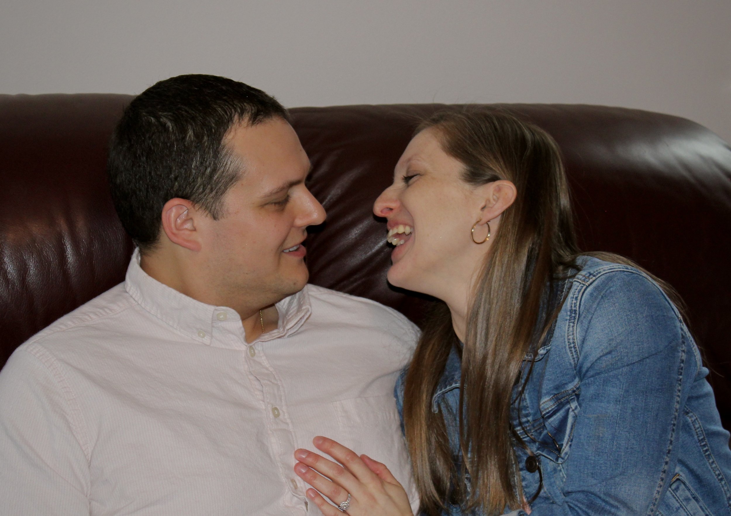 A man and woman are sitting close together on a brown leather couch, touching foreheads and smiling at each other in a cozy, intimate moment.