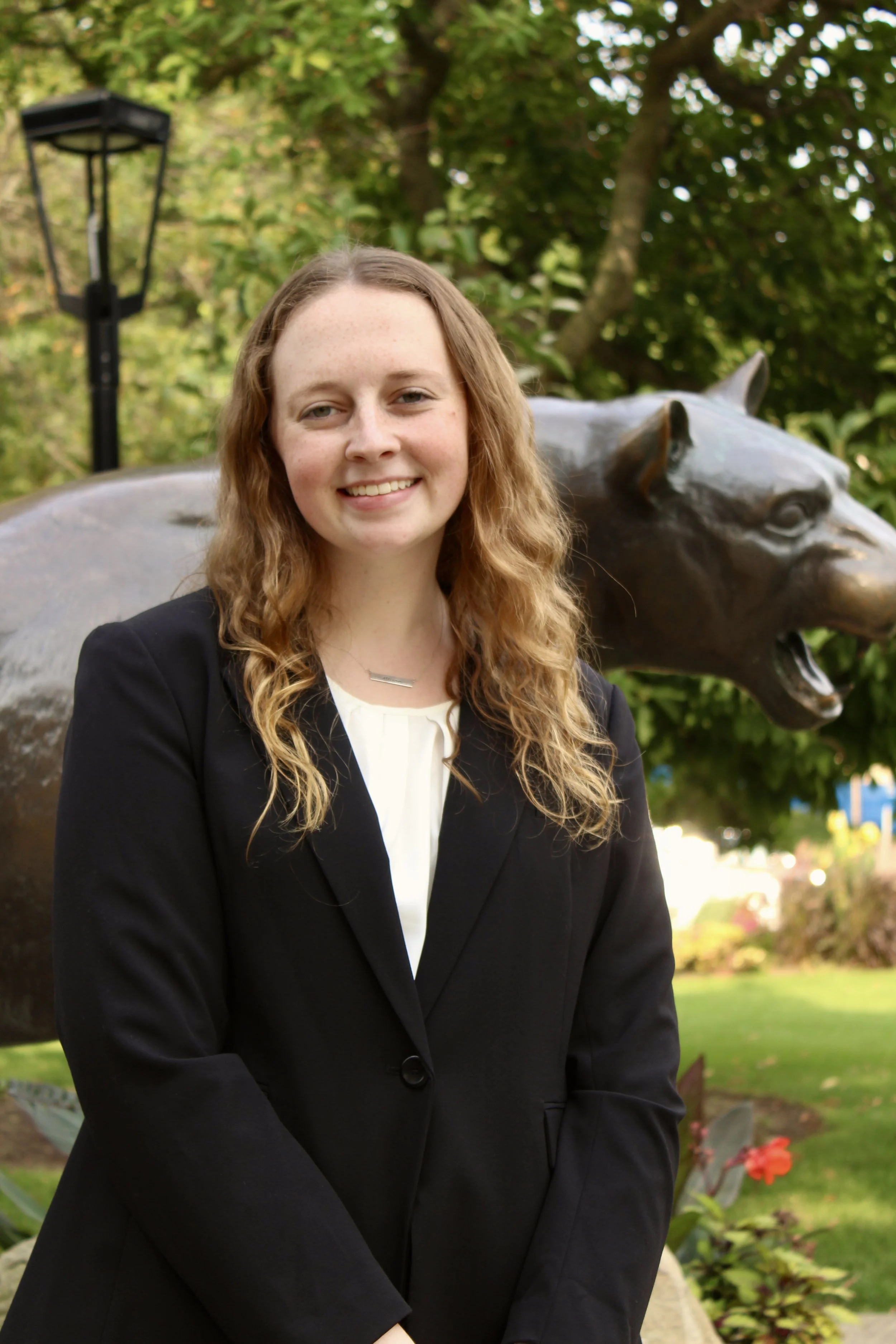 Young woman with long curly hair smiling, standing outdoors in front of a bronze sculpture of a lion's head, with trees and a park lamp in the background.