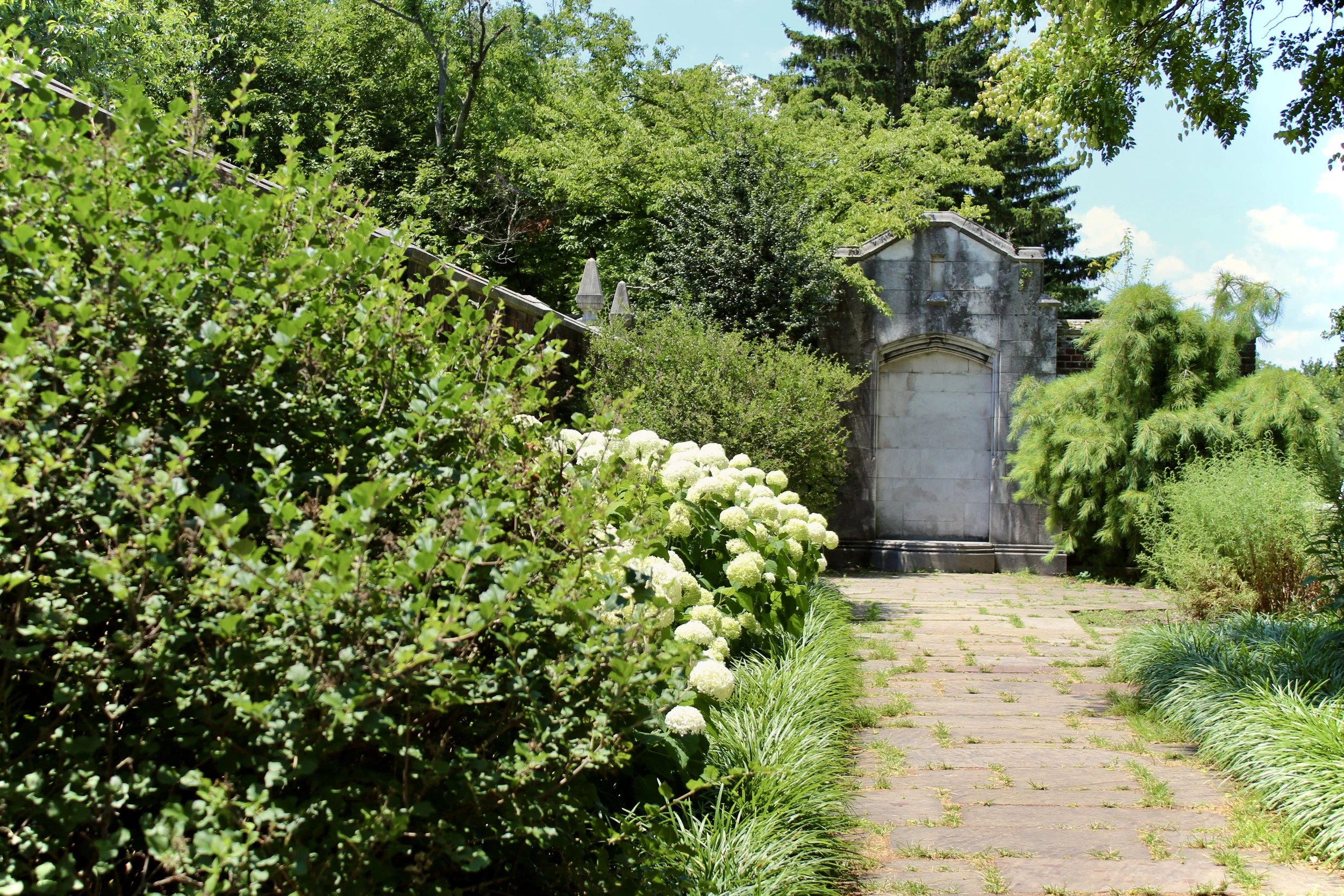 A pathway through a lush, green garden with dense bushes, white flowers, and trees leading to a stone structure with a gate and a small archway, under a partly cloudy sky.