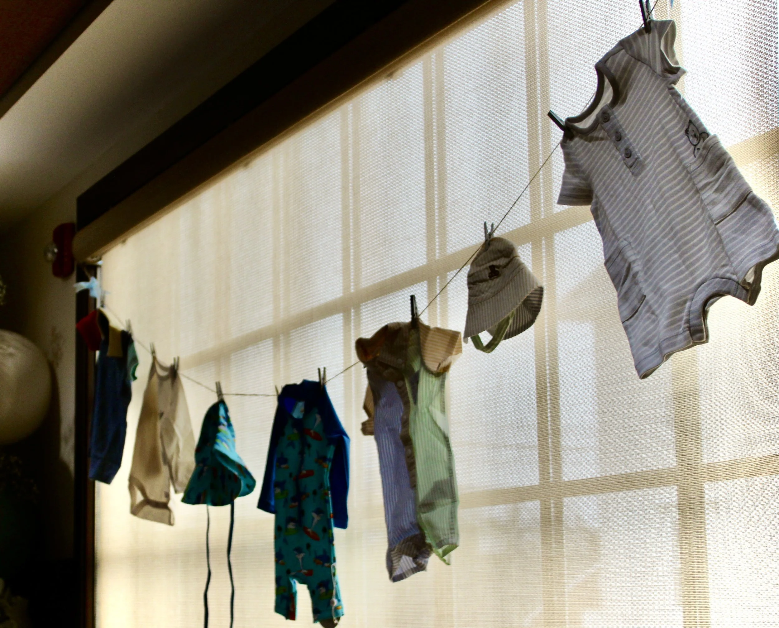 Children's clothes hanging on a string indoors, with a window and blinds in the background.