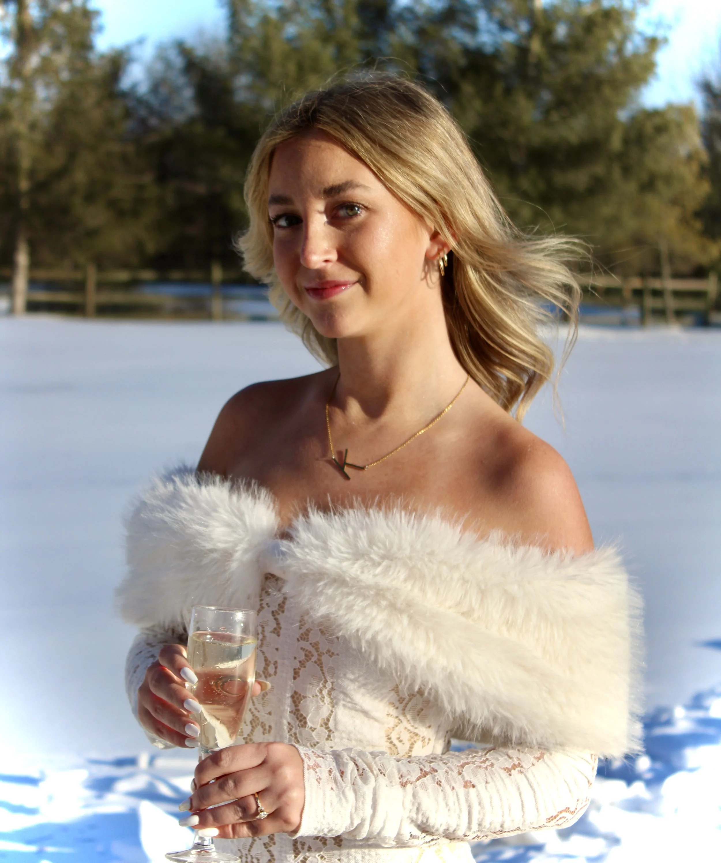 A woman wearing a white lace dress and a white fur stole holding a glass of champagne in an outdoor snowy setting, with trees and blue sky in the background.