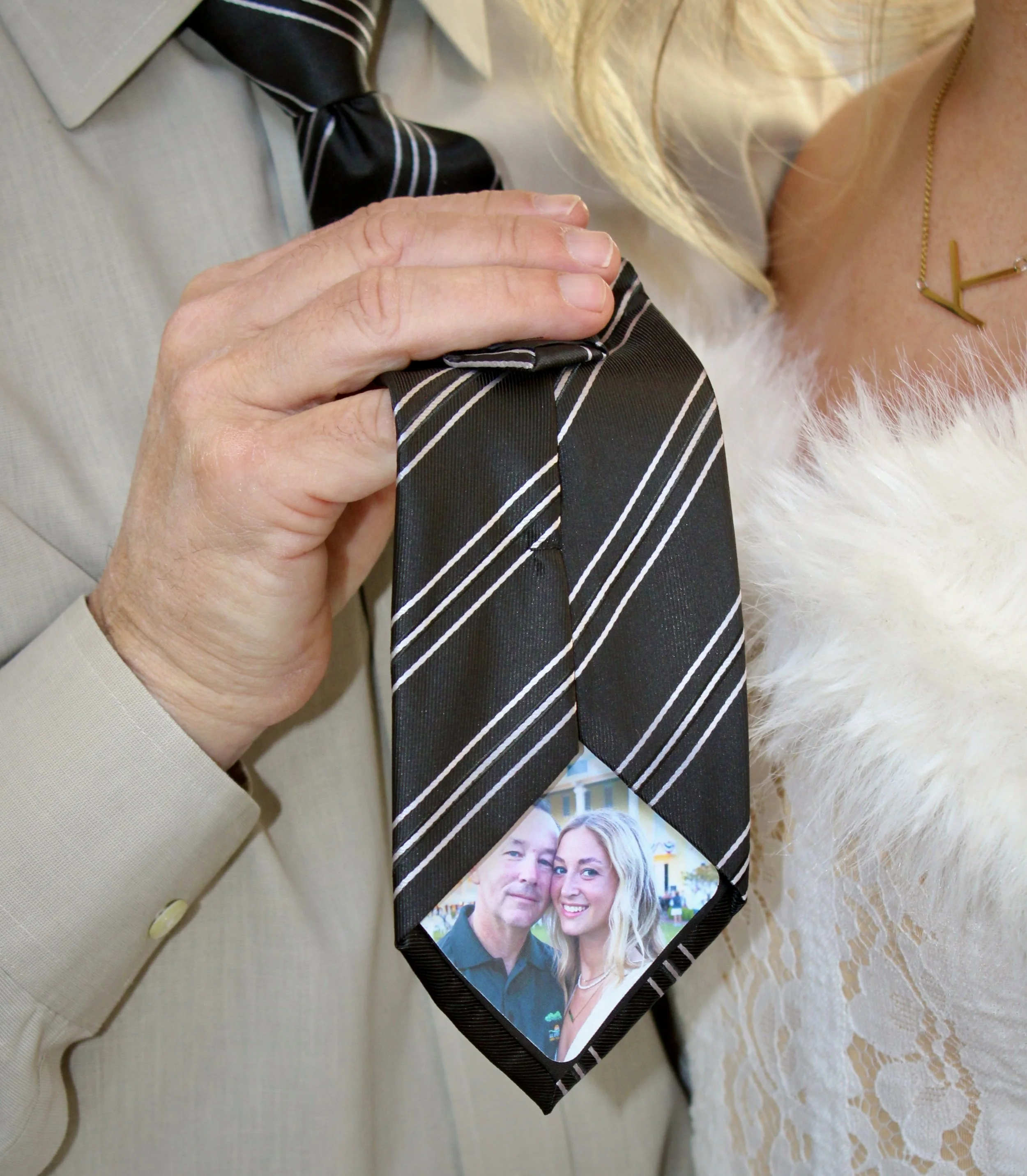 Close-up of a hand holding a black photo frame with a picture of a smiling man and woman, placed on a beige table with a lace tablecloth. Part of a person in a beige suit and striped tie is visible nearby.