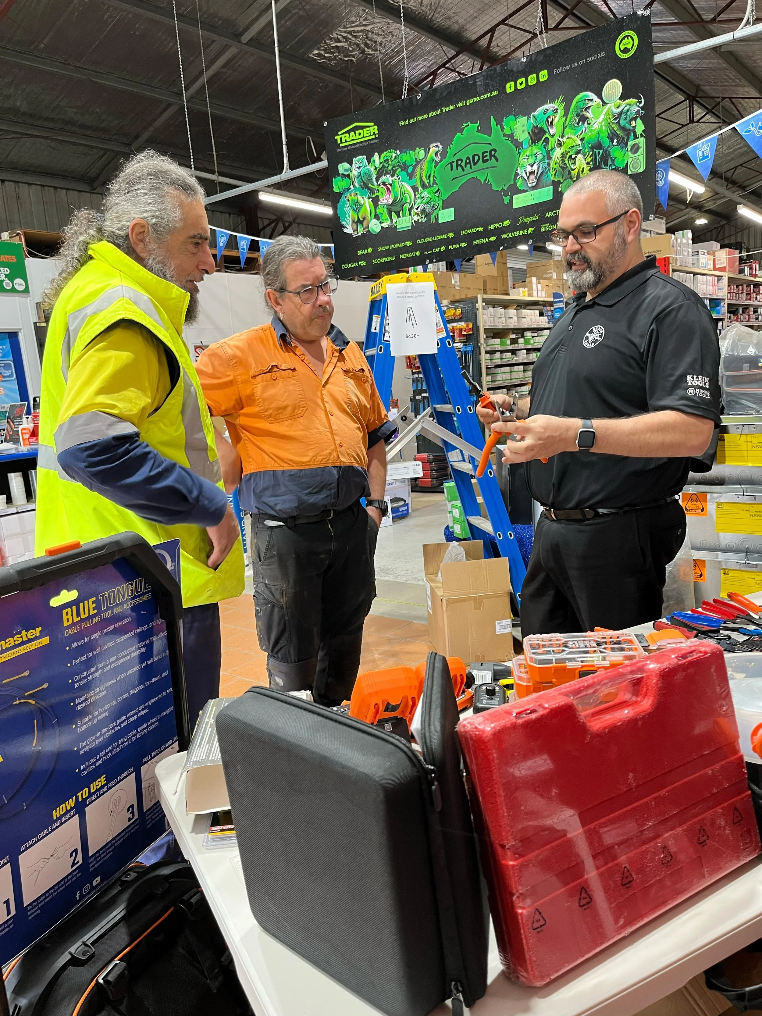 Three men standing and talking inside a hardware store, with tools and equipment on a nearby table, and a large sign overhead displaying various cartoon animals.