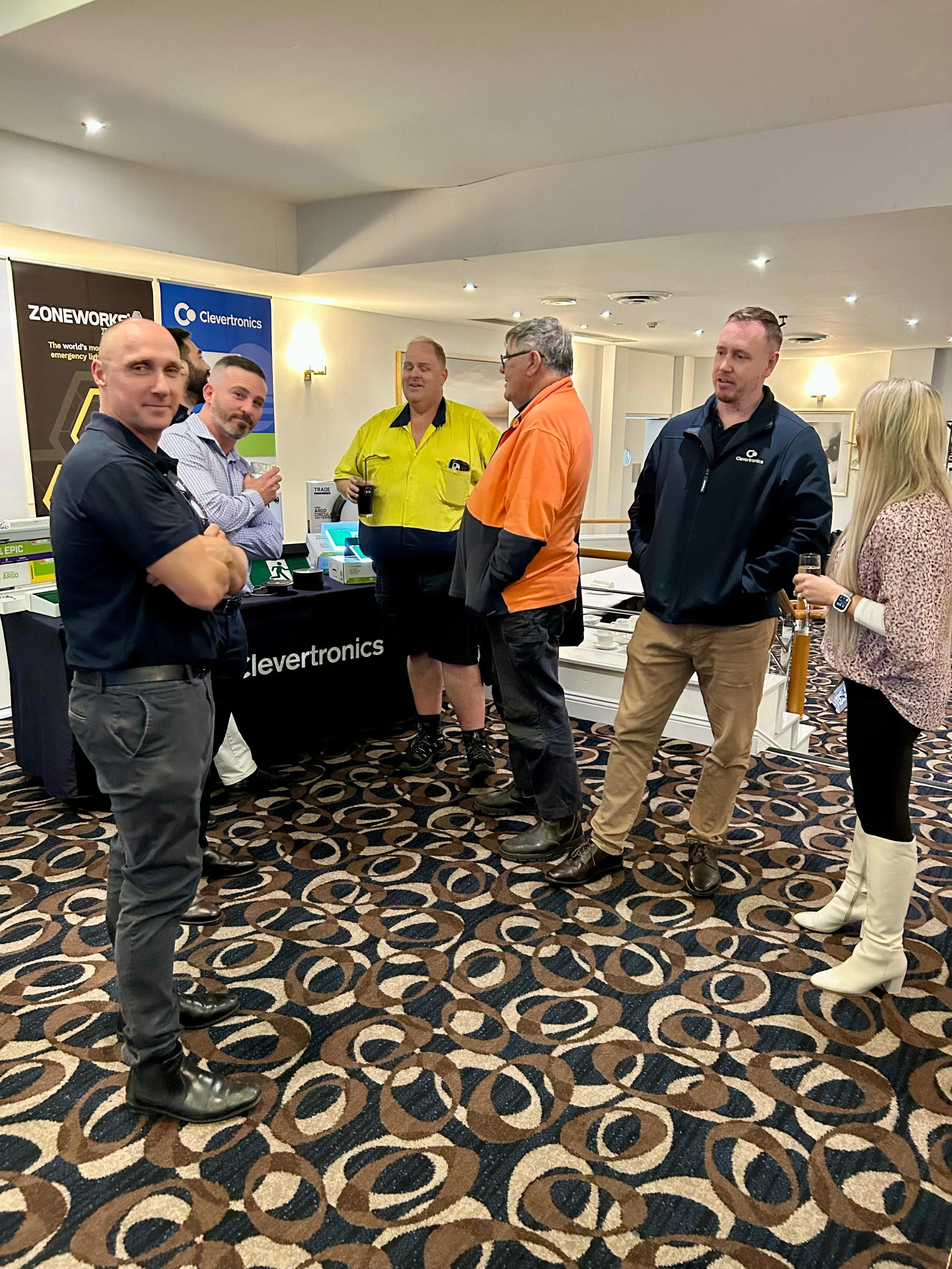 Group of six people standing and talking at a trade show or indoor event, with promotional banners and tables in the background.