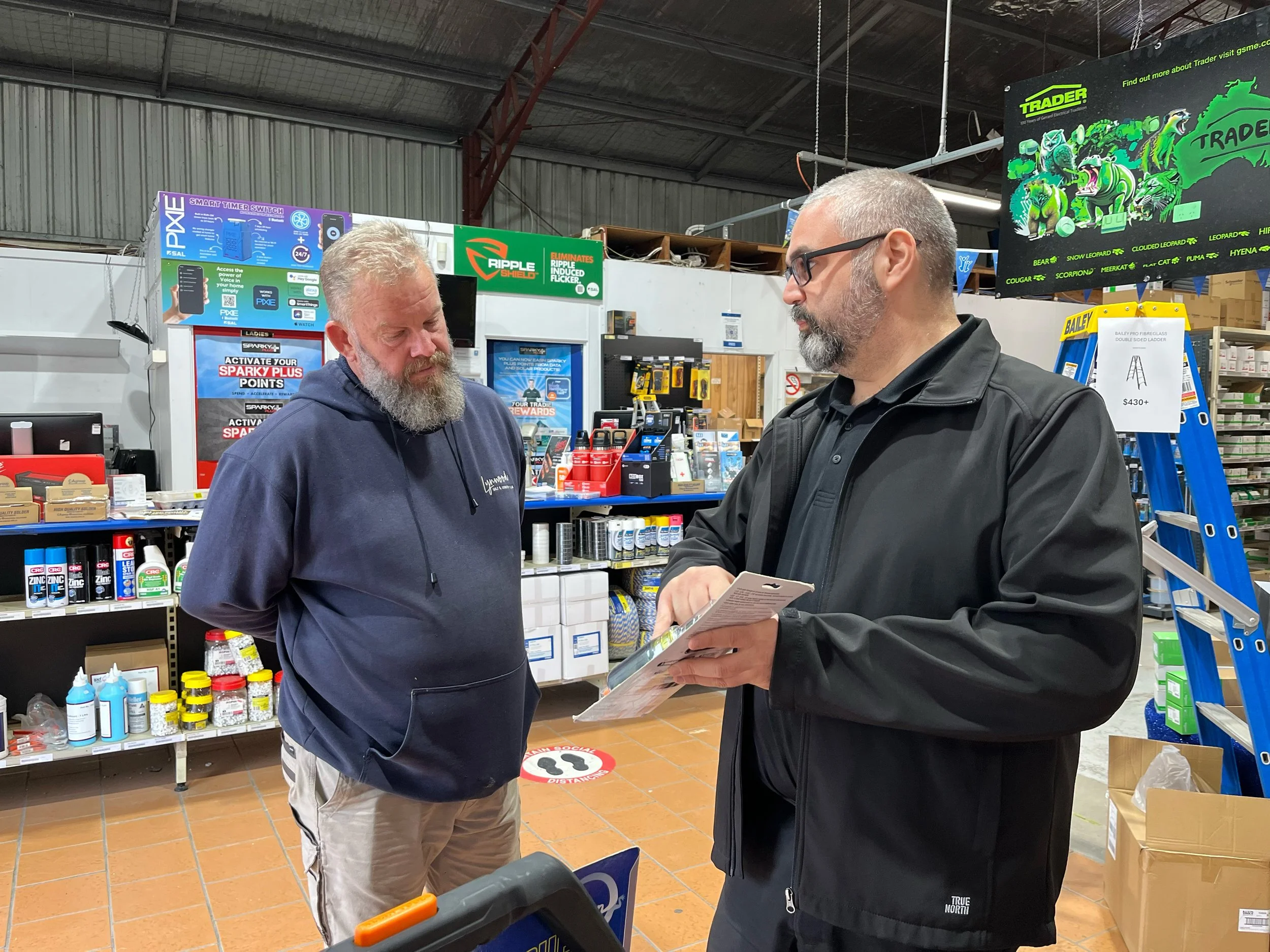 Two men are standing inside a hardware store. The man on the left has a gray beard and is wearing a navy hoodie and beige pants. The man on the right has a beard, glasses, and is wearing a black jacket and holding a booklet. The store has various sup