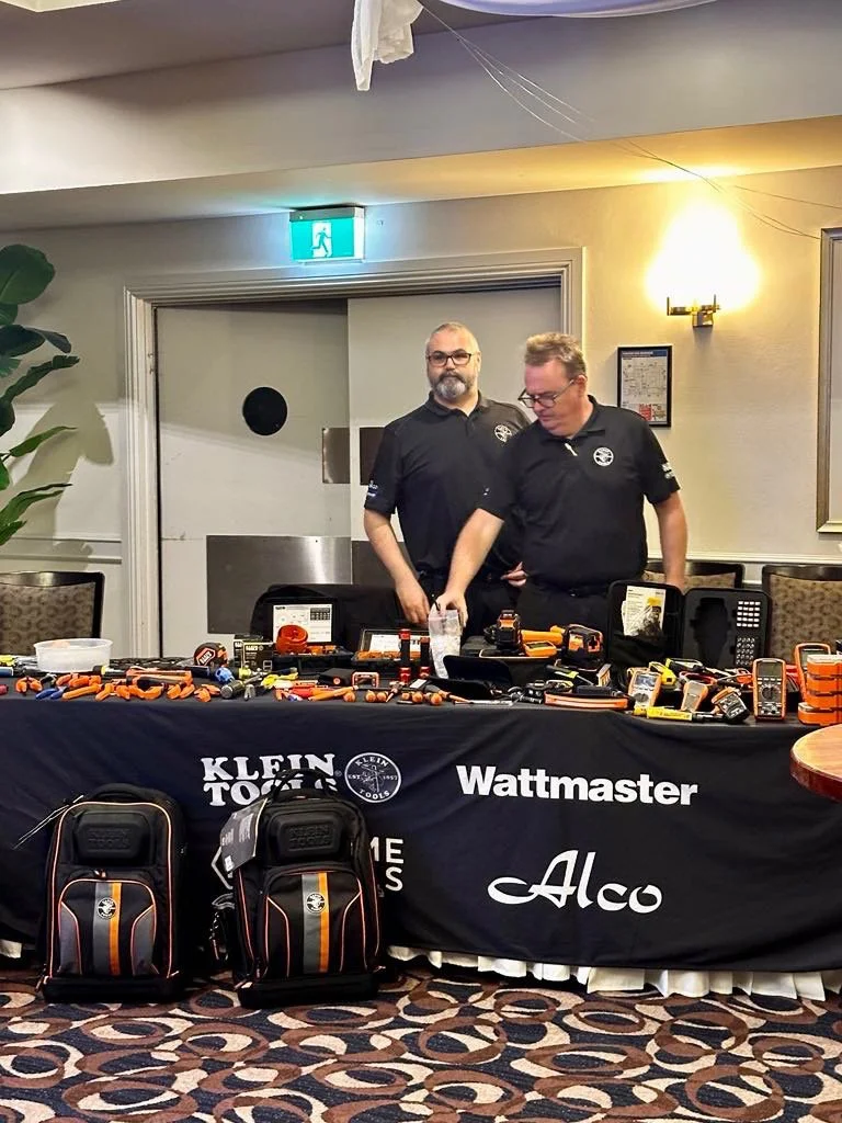 Two men in black shirts standing behind a table covered with various small electronic gadgets and tools, with backpacks on the floor in front of the table, at an indoor event or conference.