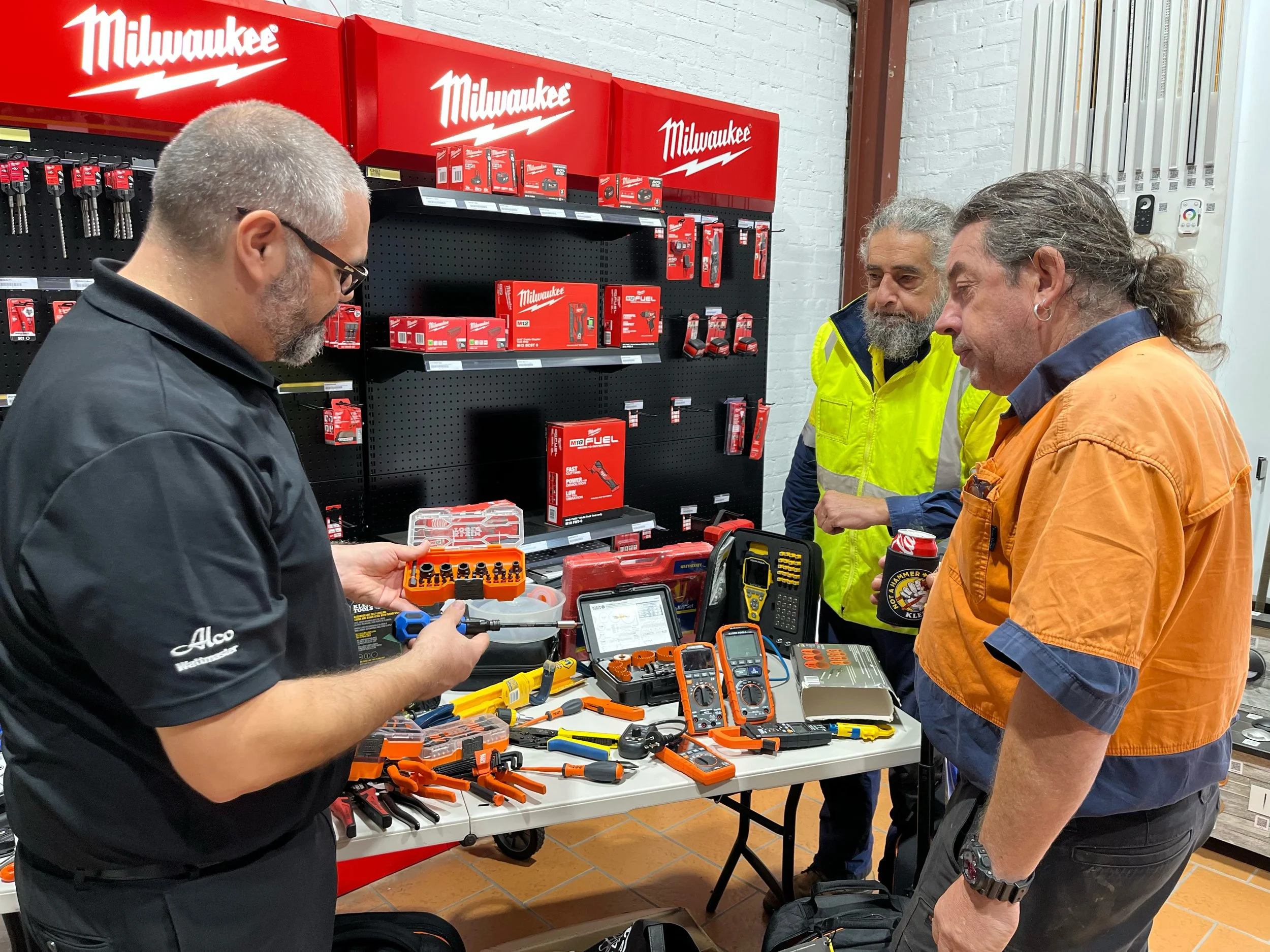 Three men at a Milwaukee tool display checking electrical testing and measurement equipment while the man on the left holds a tool in his hands.