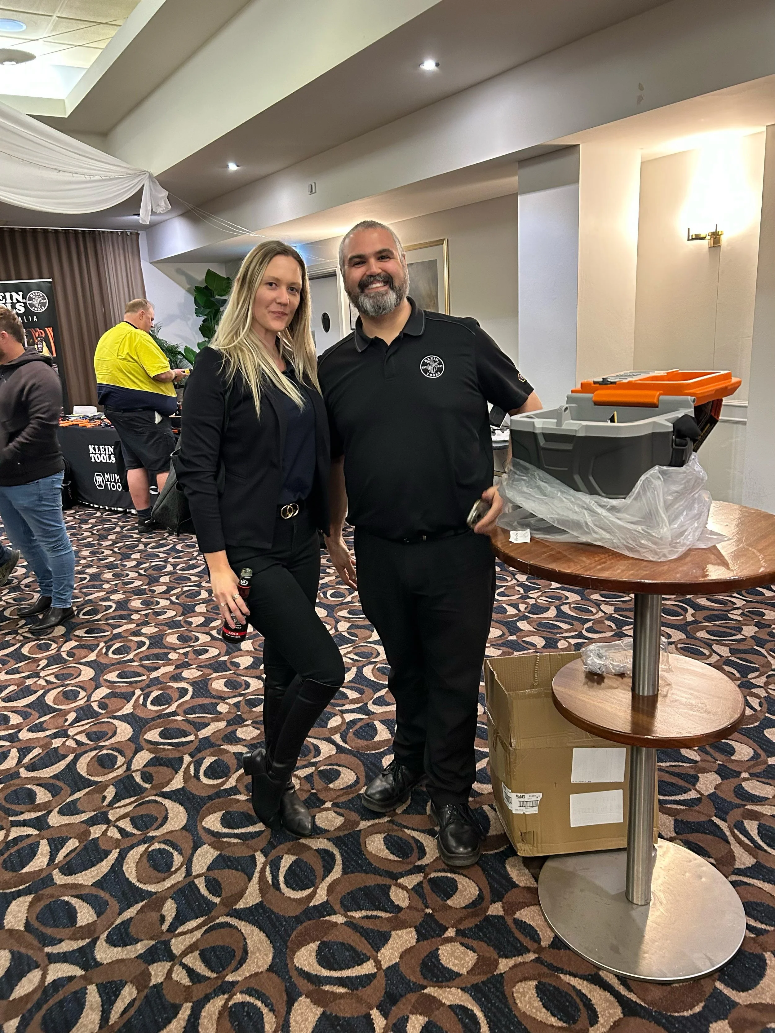 A woman and a man standing together in a room, smiling at the camera. The woman is holding a beer bottle, and the man has his arm around her. There are people and tables in the background, and the floor has a patterned carpet.