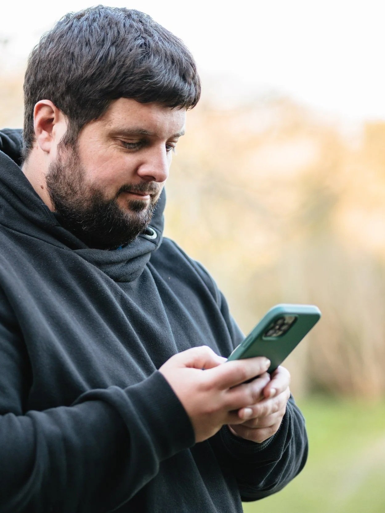 A man with a beard wearing a black hoodie looks down at his smartphone outdoors, with a blurred background of trees.