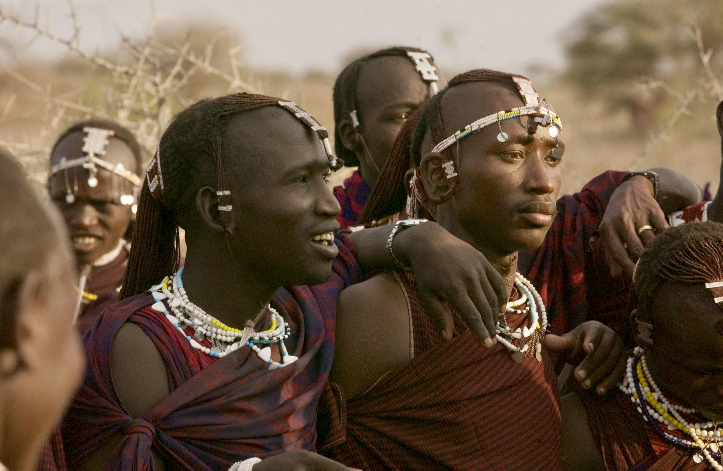 Group of Maasai people wearing traditional jewelry and clothing, sitting outdoors.