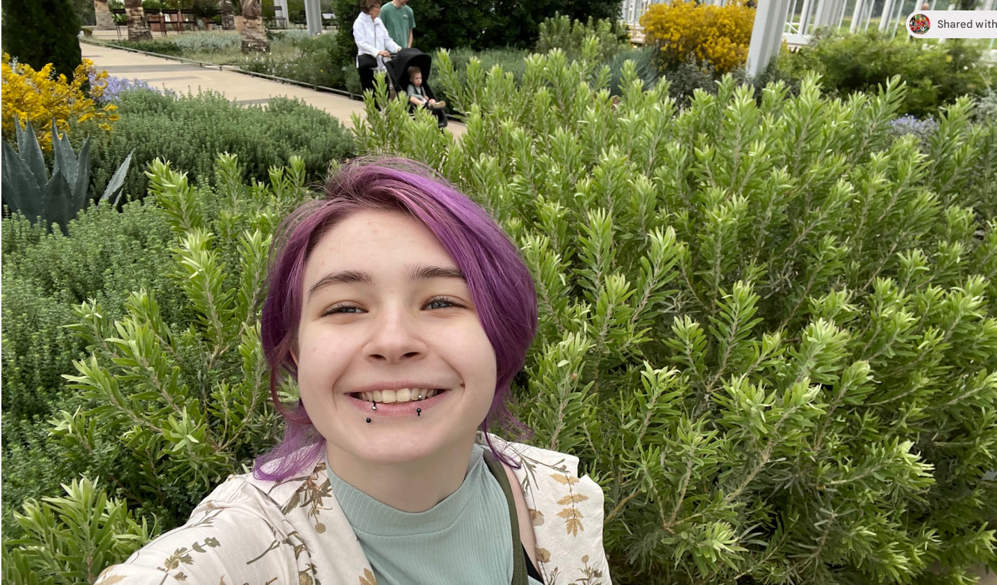 Young woman with purple hair smiling outdoors in front of green bushes, in a garden with other people and plants in background.
