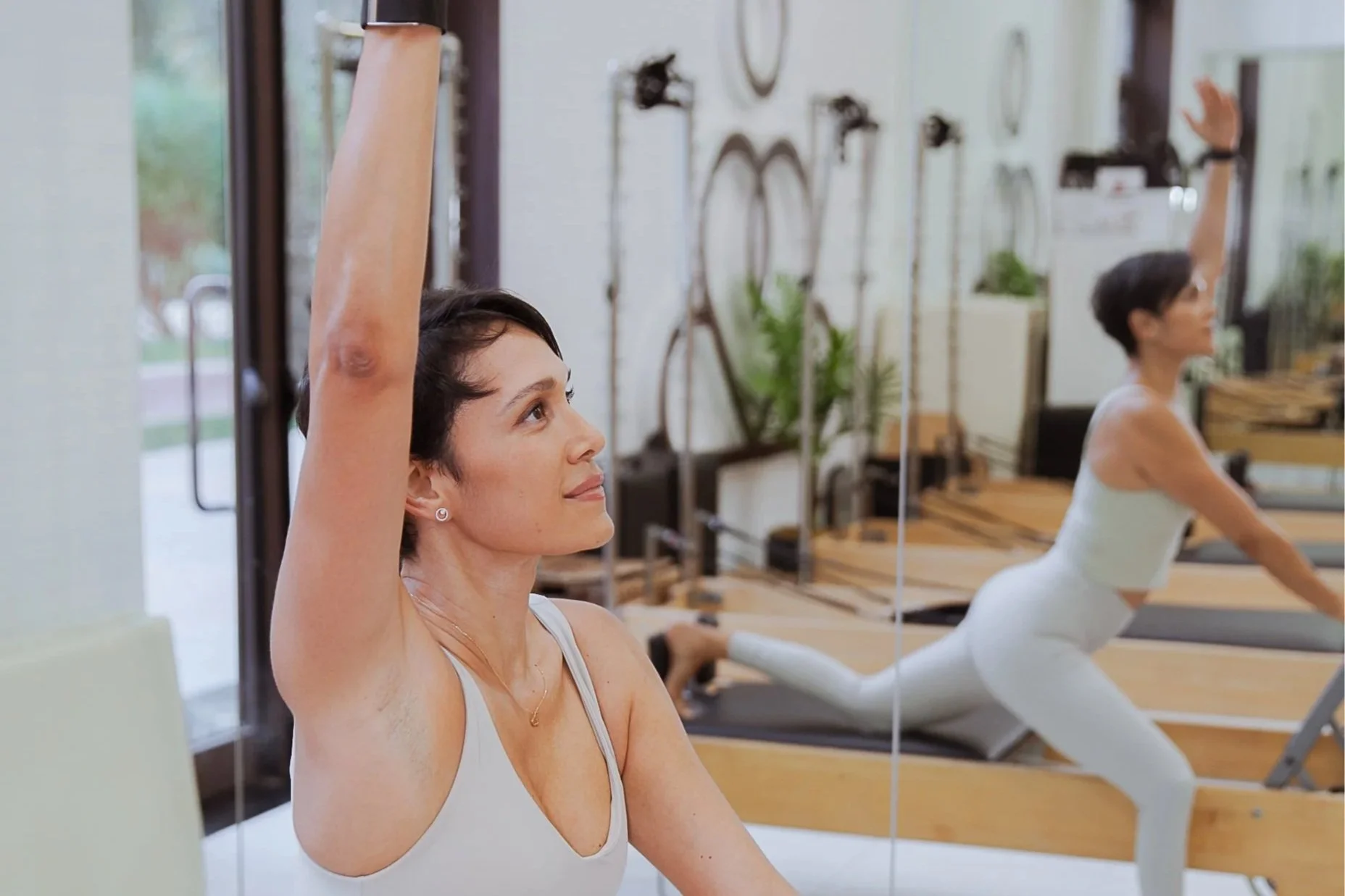 Paula Urive in a white tank top practicing Pilates by stretching her arm upward in front of a mirror at a fitness studio.