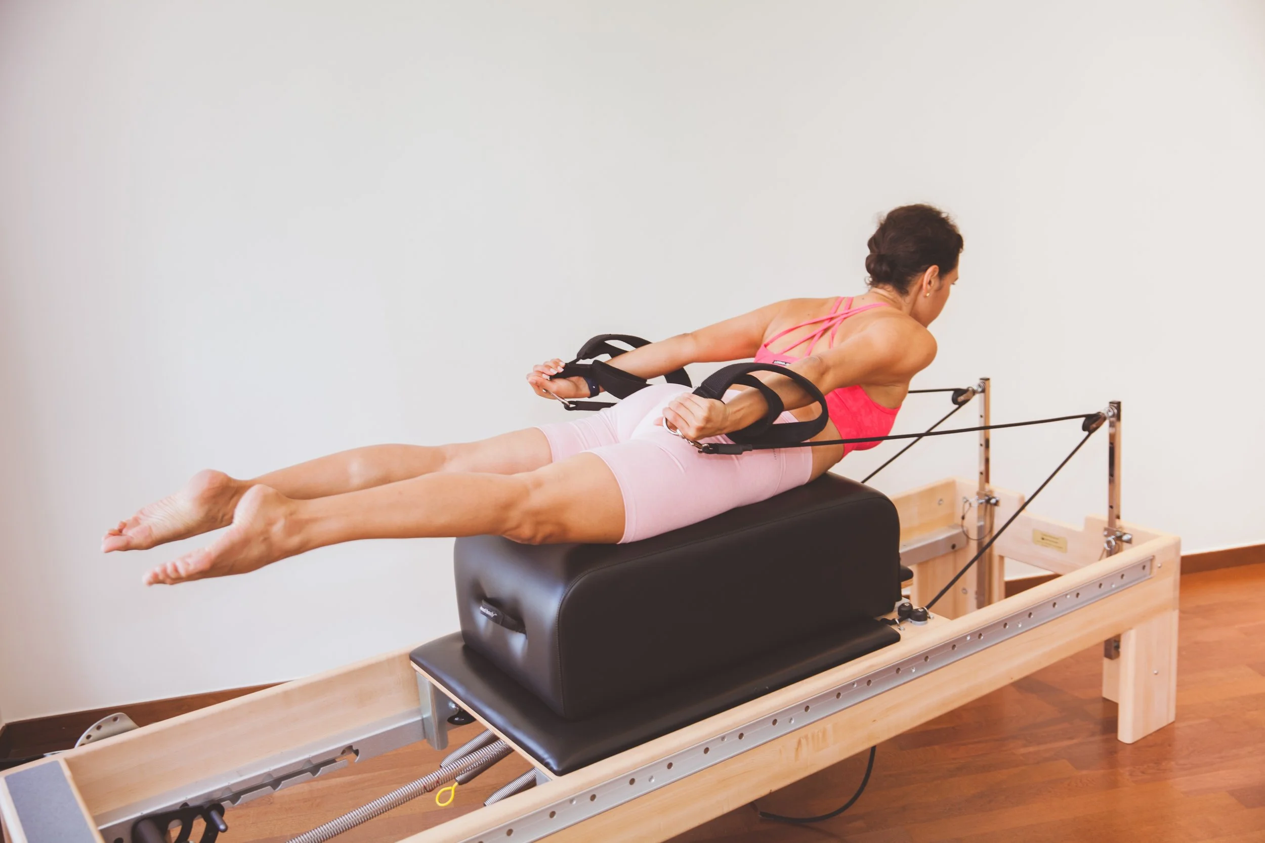 Pilates instructor Paula Uribe in pink workout attire lying face down on a Pilates reformer machine with a box, performing an exercise with black resistance straps attached to the machine.