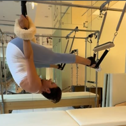 Paula Uribe in a white shirt and light pants hanging upside down on a Pilates Cadillac, holding onto the handles with her feet suspended in the air.