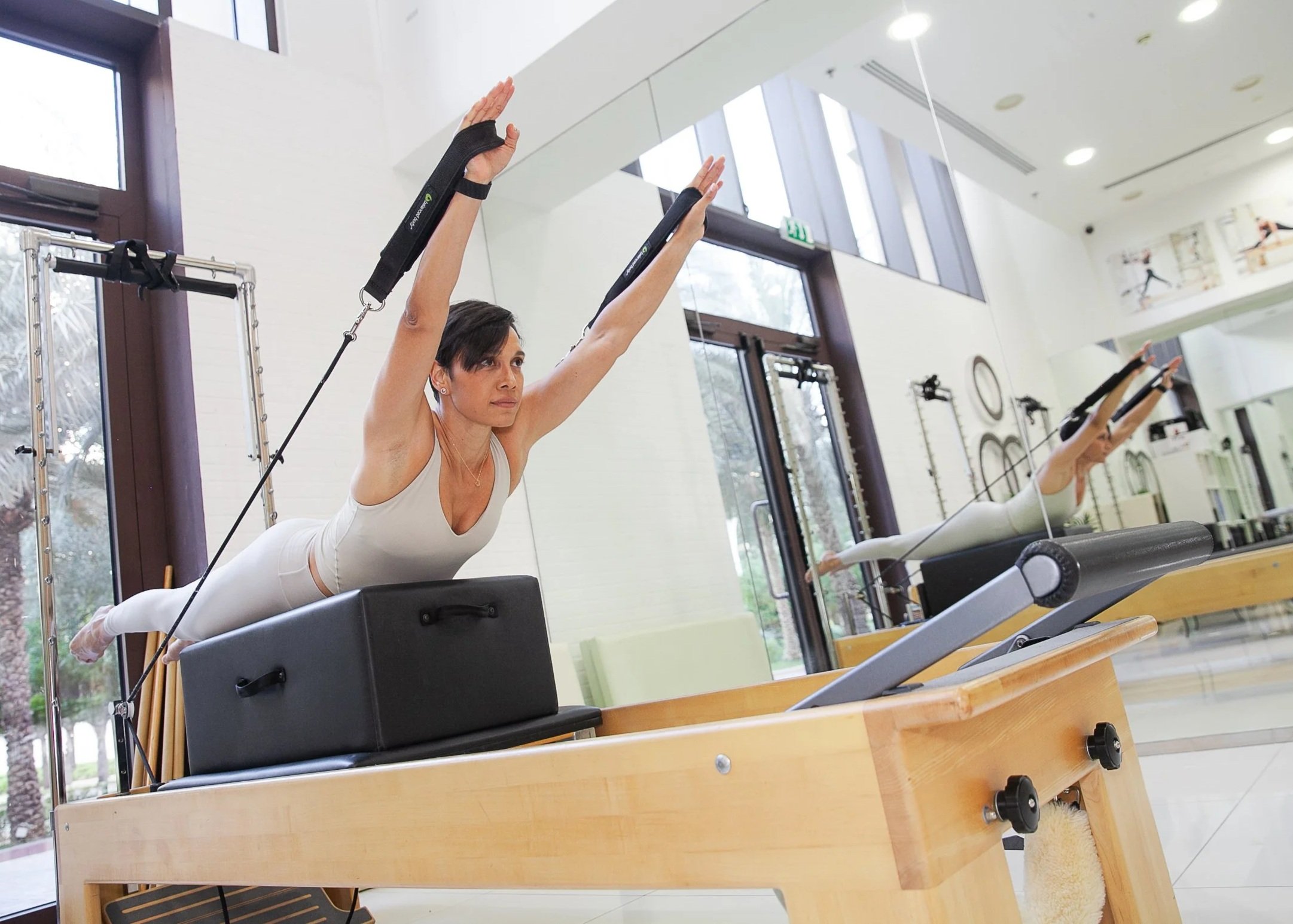 Paula Uribe practicing Pilates on a reformer machine in a bright fitness studio with large windows and mirror