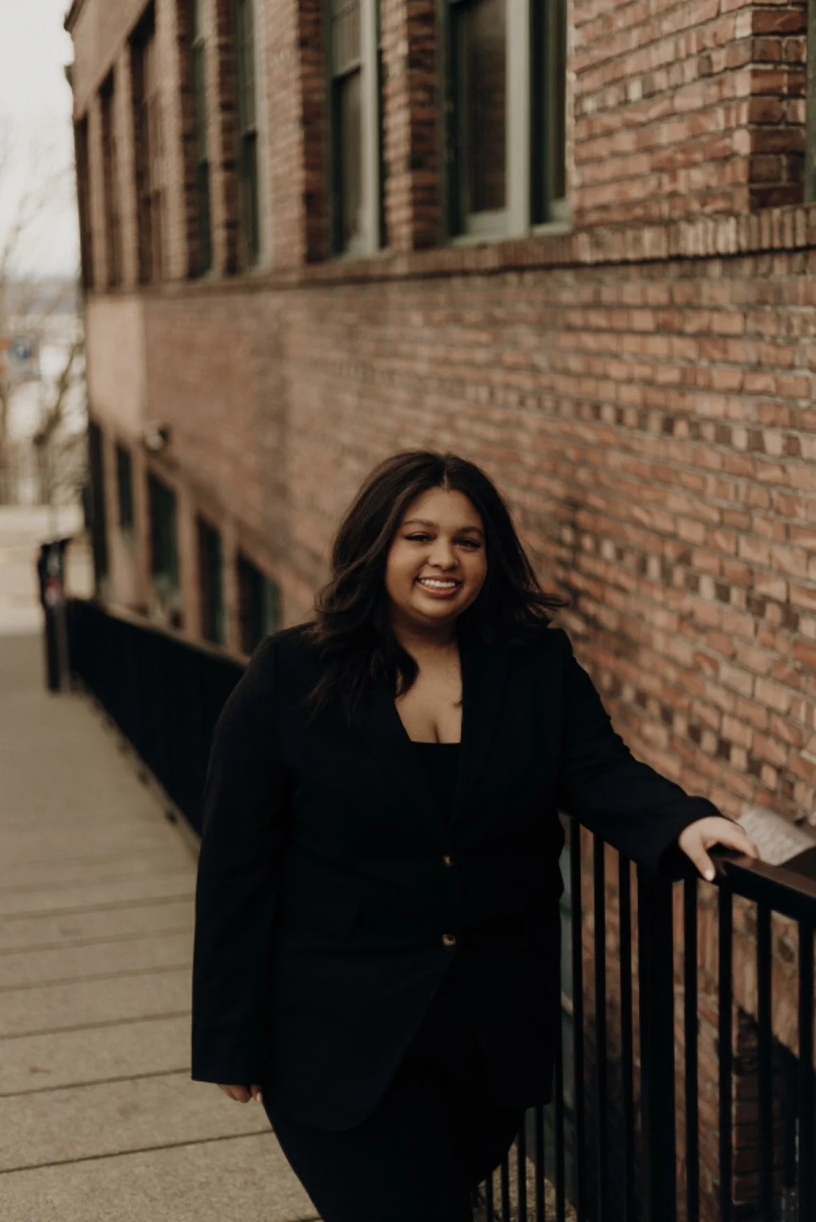 A woman with dark hair smiling, wearing a black blazer, standing on a sidewalk next to a brick building with green window frames.