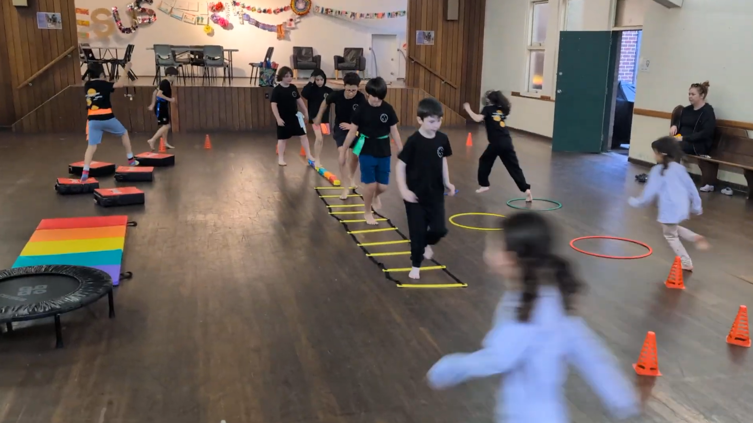 Children participating in indoor obstacle course activities, practising agility, teamwork, coordination, and developing fitness while being supervised by an adult.