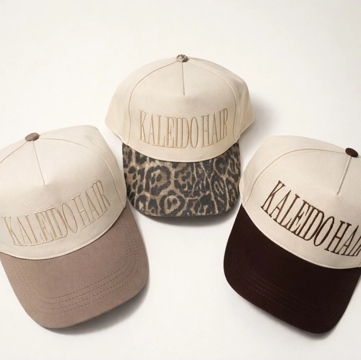 Three baseball caps with "KALEIDOHAR" embroidered on the front, featuring different colored and patterned brims: one with a beige brim, another with a leopard print brim, and the third with a dark brown brim.