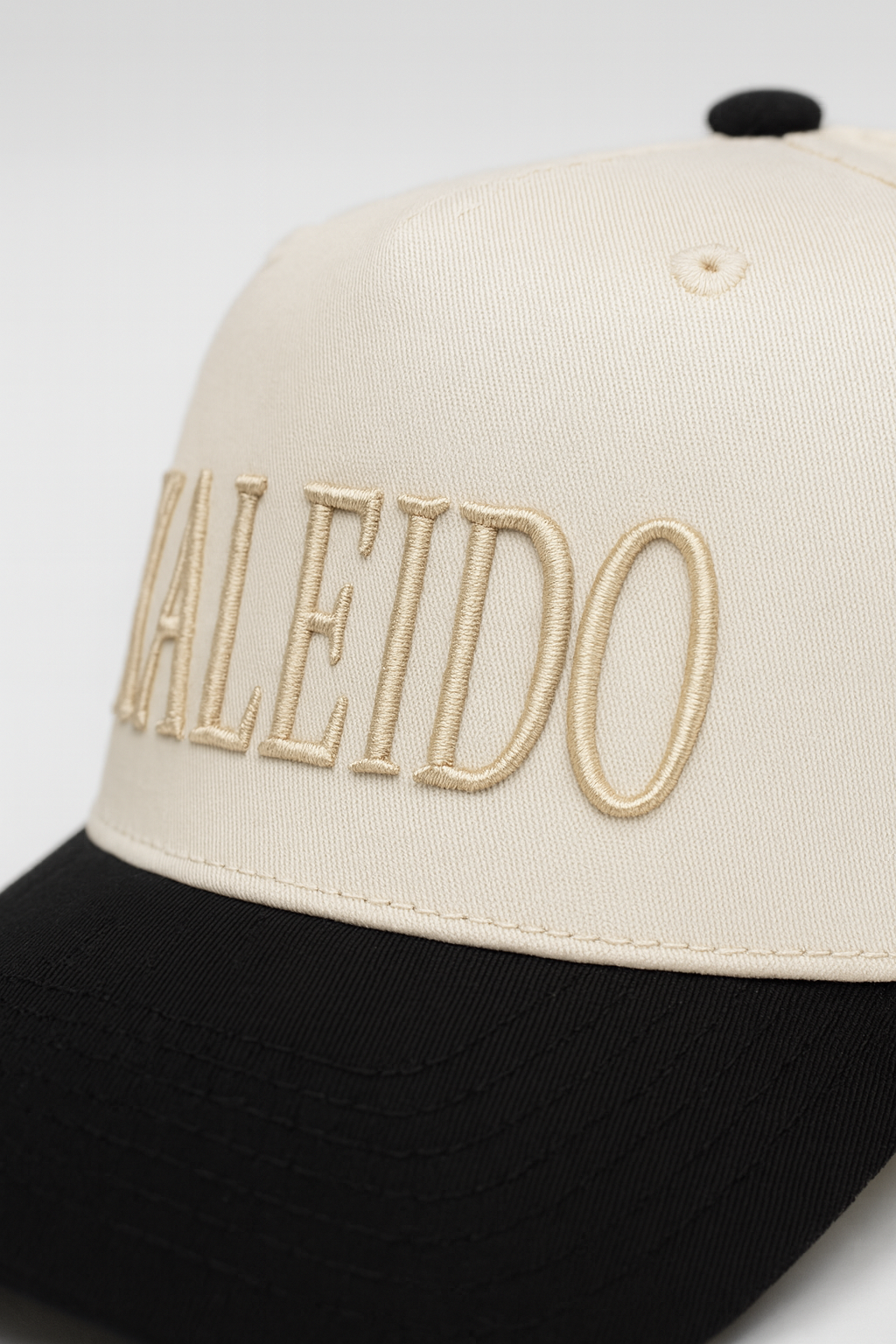 A two-tone baseball cap with a cream crown and black bill, featuring 'ALHEIDO' embroidered on the front in beige thread.
