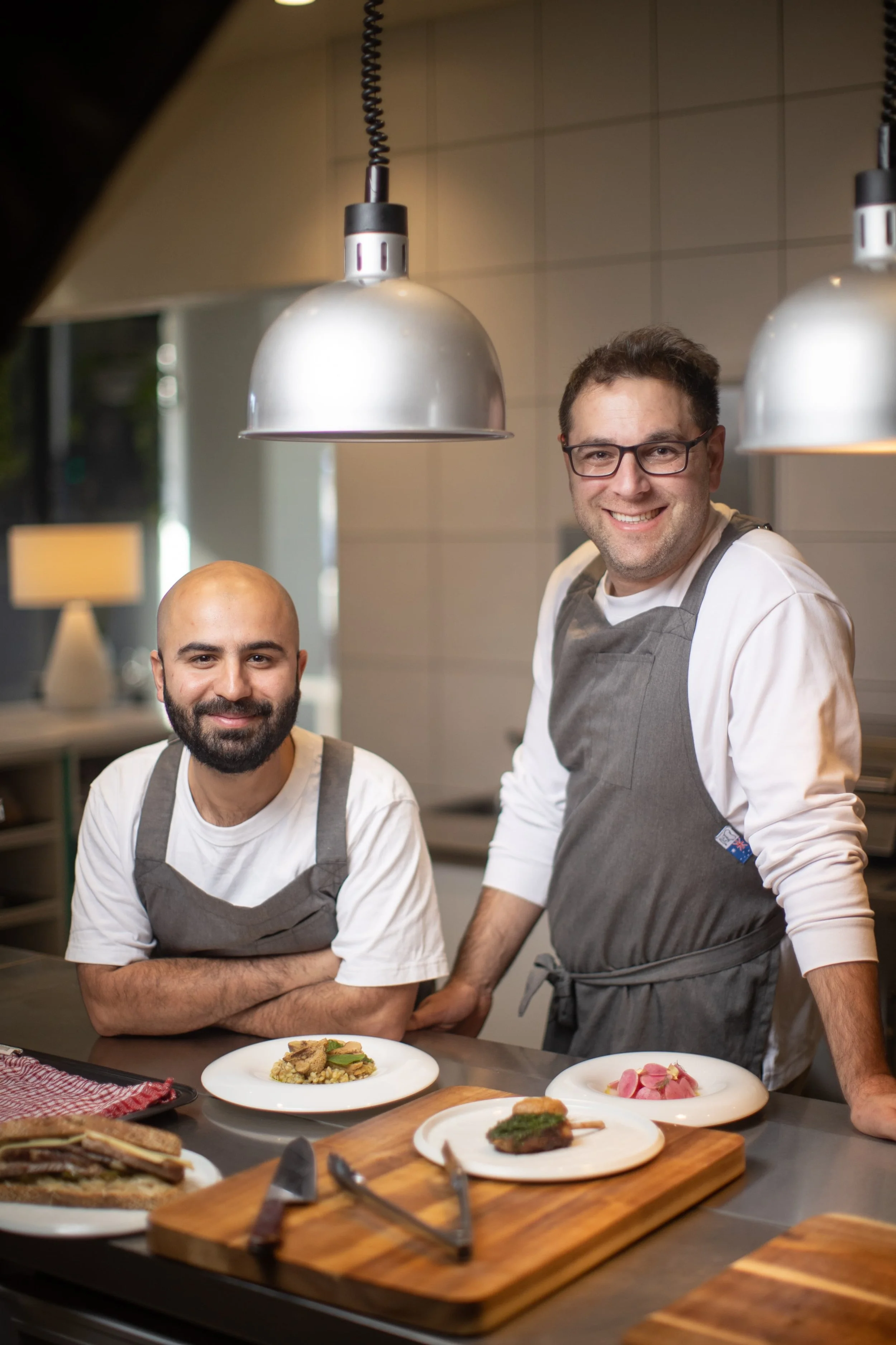 Two male chefs in aprons in a professional kitchen, smiling at the camera with plates of food in front of them.