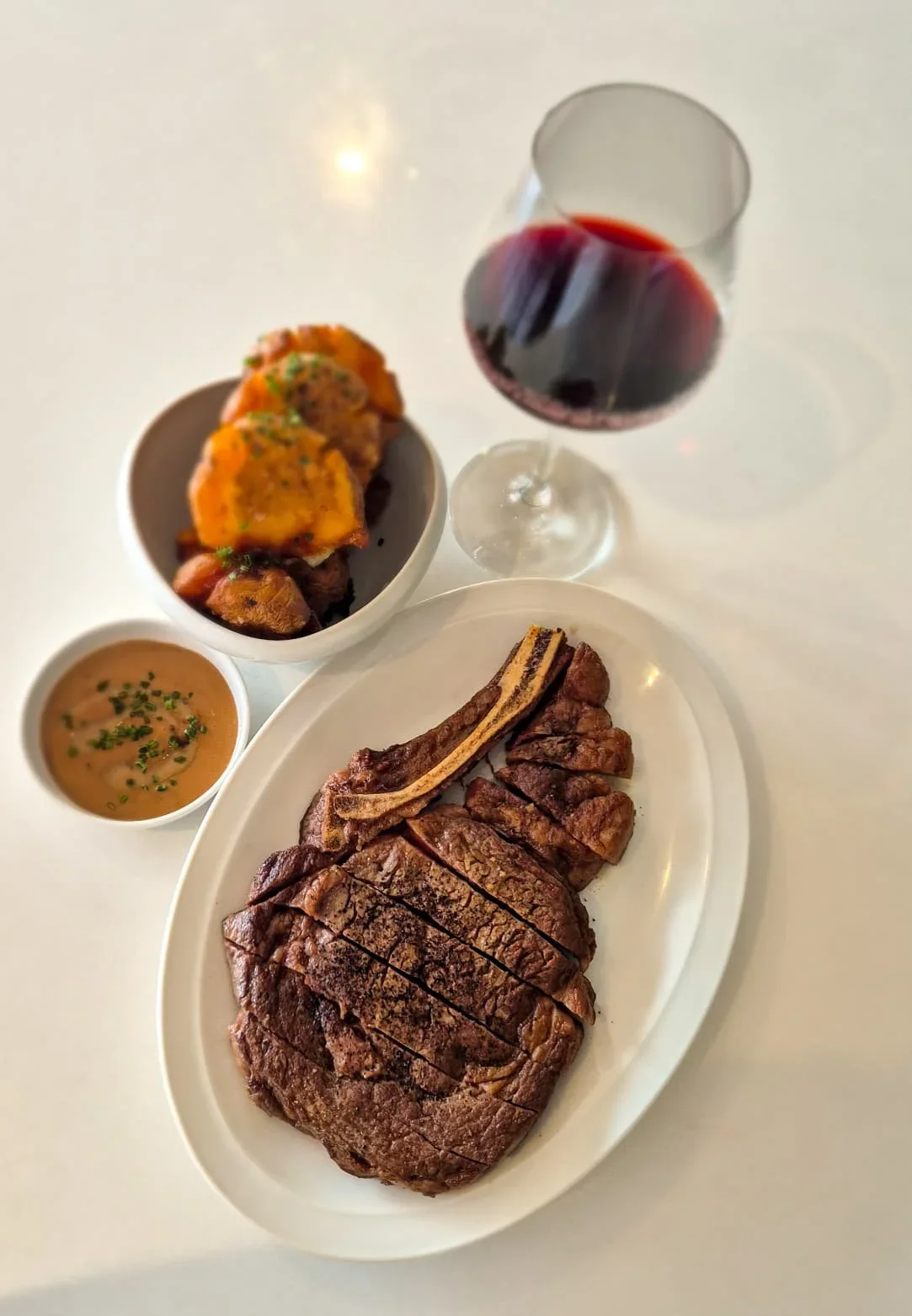 A plate with sliced ribeye steak, a bone-in beef rib, a bowl of sauce, a bowl of roasted vegetables, and a glass of red wine on a white table.
