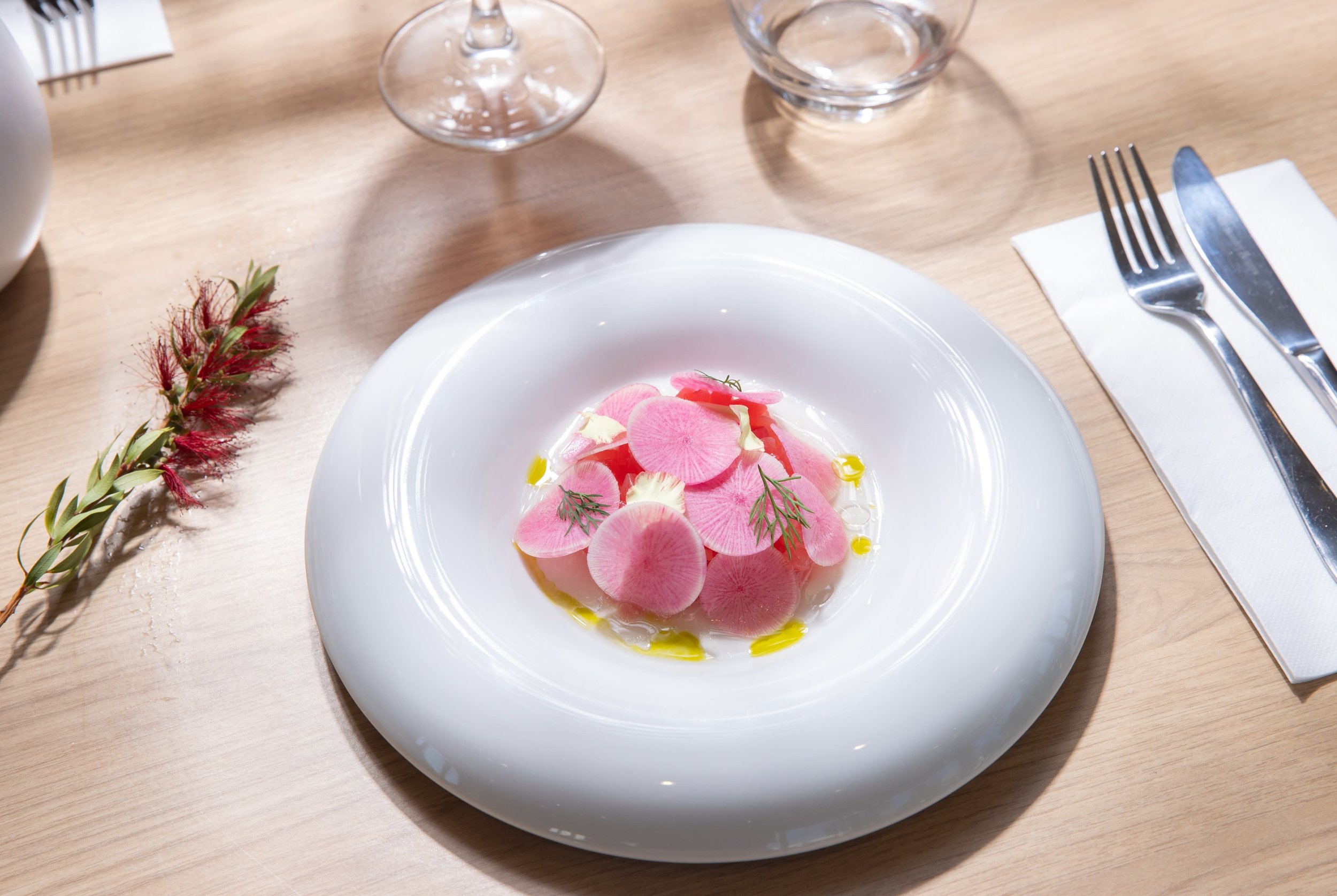 A dish of sliced pink radishes garnished with herbs, with a drizzle of olive oil, served on a white plate.